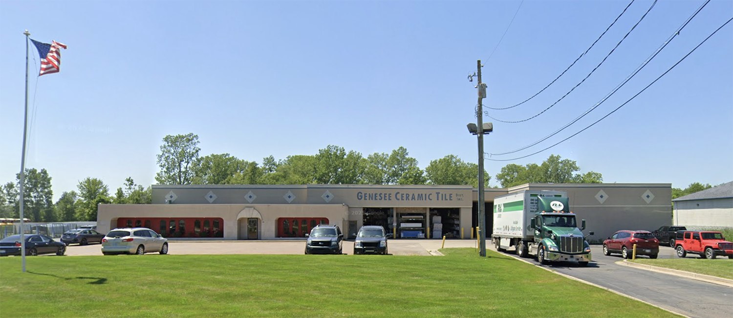 A one-story building labeled “Genesee Ceramic Tile” has cars parked in front and a truck on the right. An American flag flies on a pole, and trees are visible in the background under a clear blue sky.