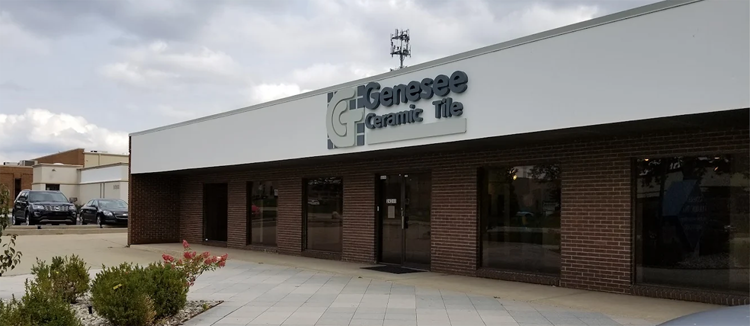 A one-story building with a sign reading Genesee Ceramic Tile above dark tinted windows; cars are parked to the left and plants line the sidewalk in front.
