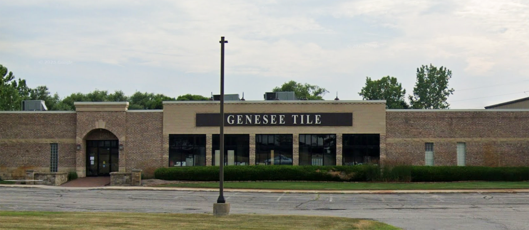 A brick building with a sign reading Genesee Tile above large windows, surrounded by greenery and a mostly empty parking lot in front.