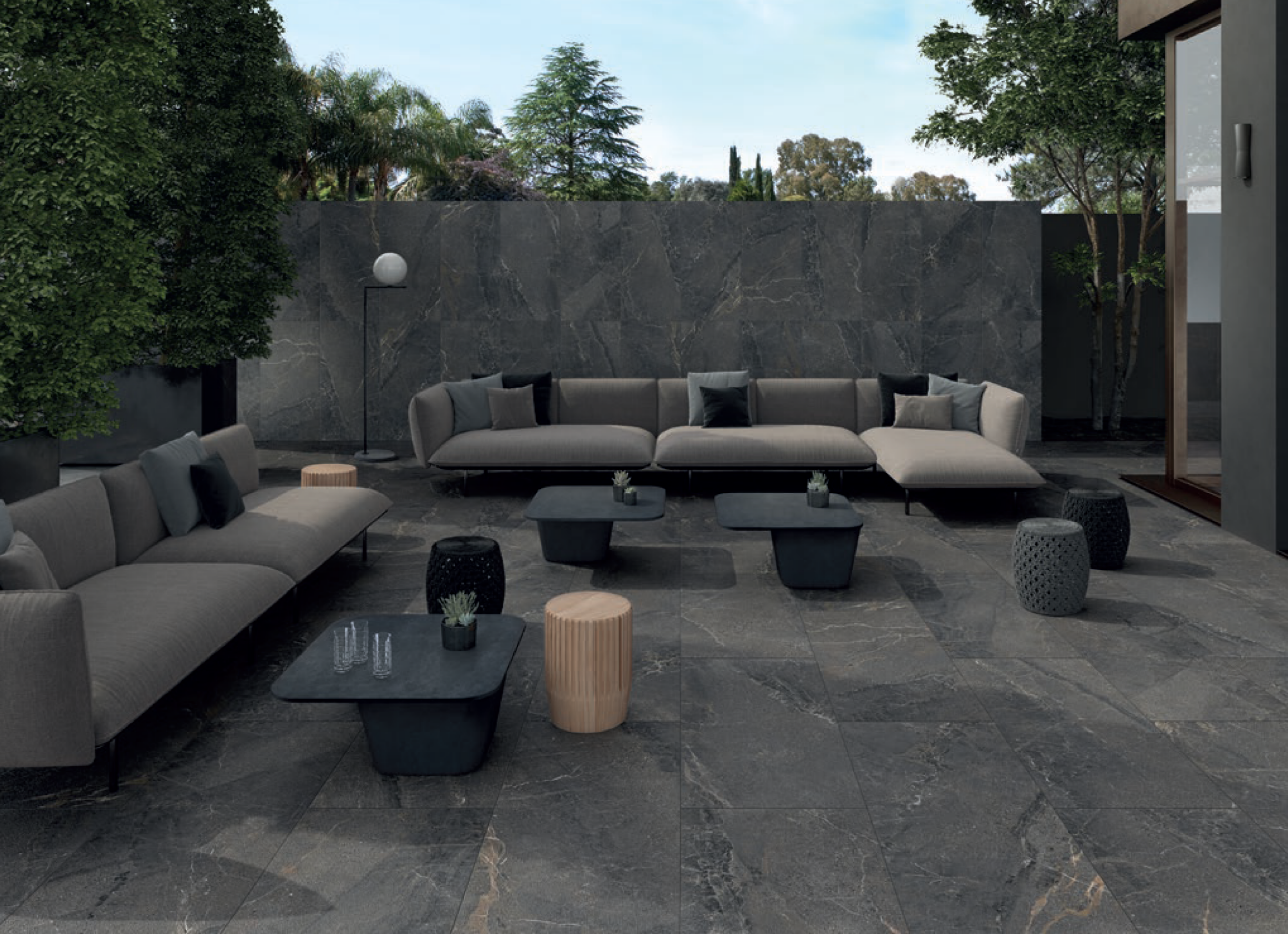 Modern outdoor patio with gray stone tiles, gray sectional sofas, black and beige tables, small stools, and greenery along a stone wall. Glasses and a carafe are on one table. Sky and trees visible in the background.