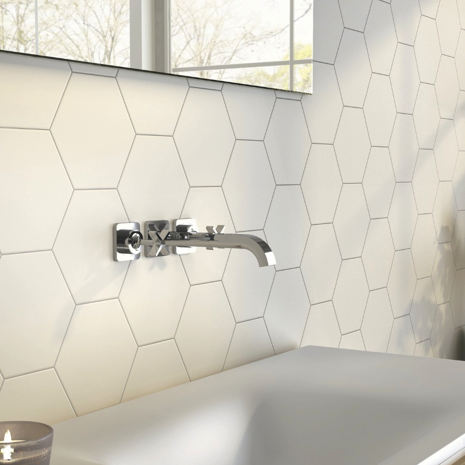 Modern bathroom with white hexagonal tile walls, a sleek chrome wall-mounted faucet, and a white sink beneath a window. A lit candle is visible in the corner.