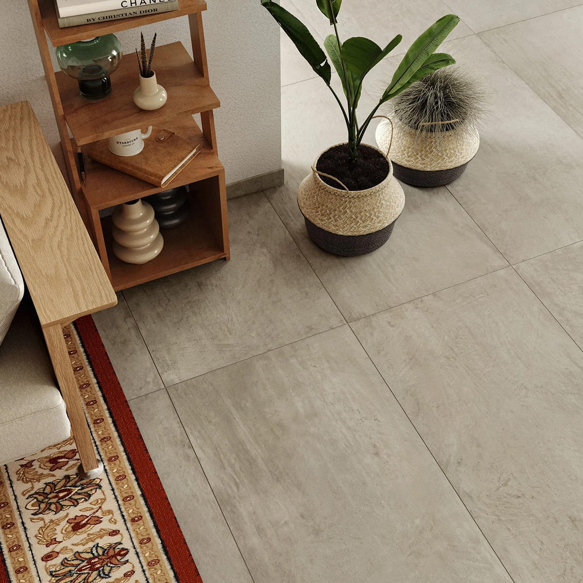 A corner of a living room with beige floor tiles, a wooden shelf holding books and a glass container, two potted plants, part of a sofa, and the Boho - Fog rug featuring red, yellow, and white designs.