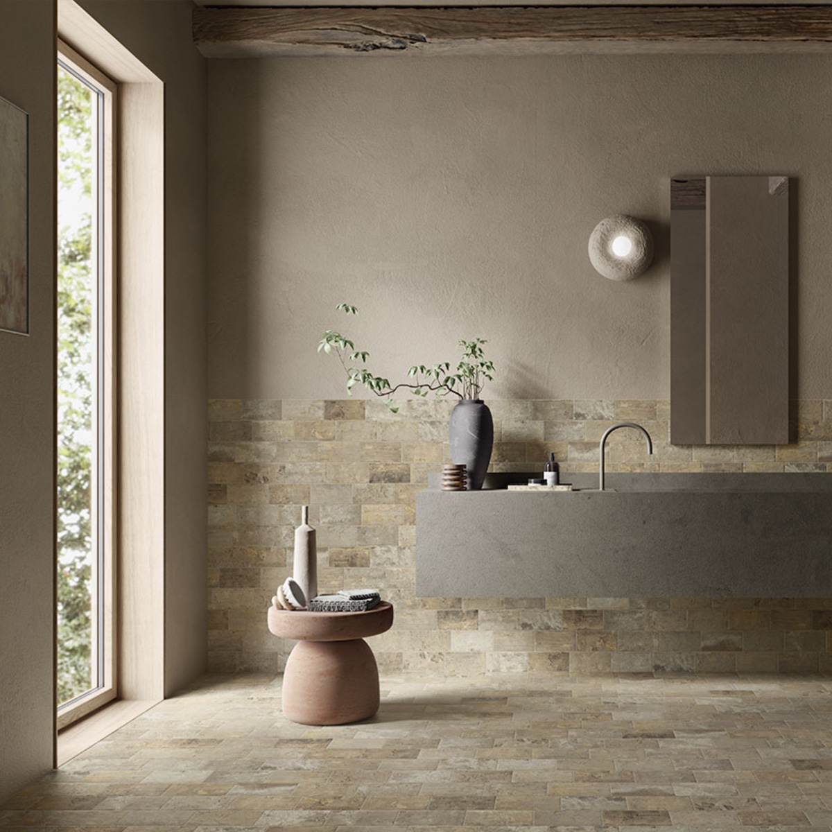 Serene minimalist bathroom with warm sandy beige stone-look brick tiles used on both the floor and as a mid-wall wainscot, paired with a floating concrete vanity, a tall dark ceramic vase with a branching plant, a round terracotta stool, and a large window overlooking trees.