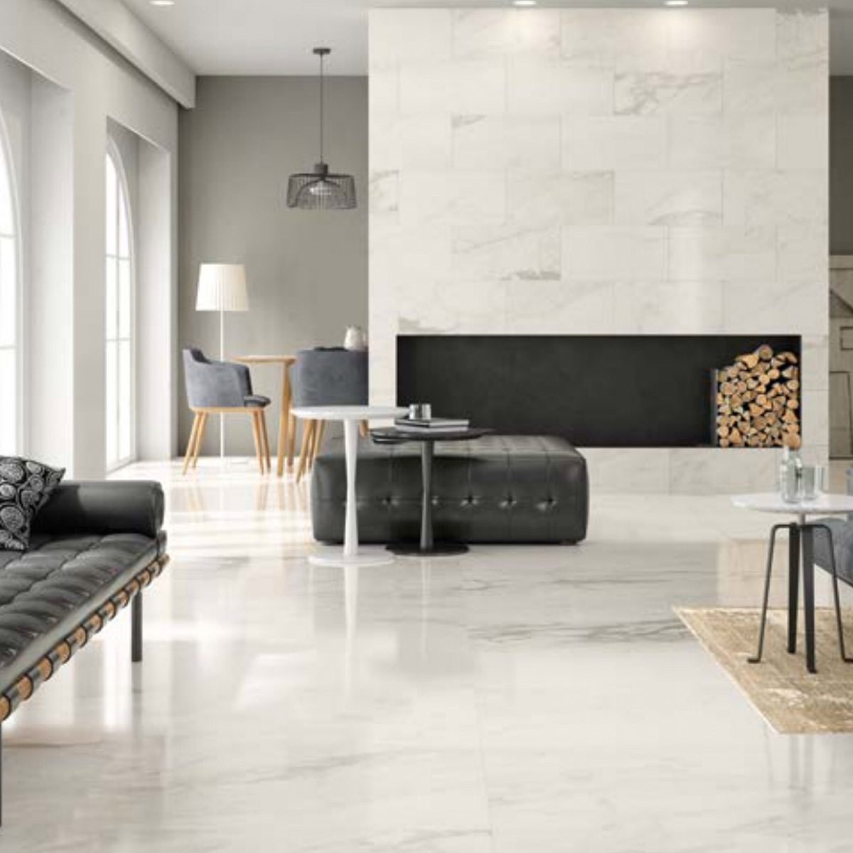 A modern living room features elegant Carrara Select 2.0 marble floors, complemented by a black sofa, round tables, gray chairs, expansive windows, and a fireplace with stacked firewood on the right. The space is bright and neatly arranged.