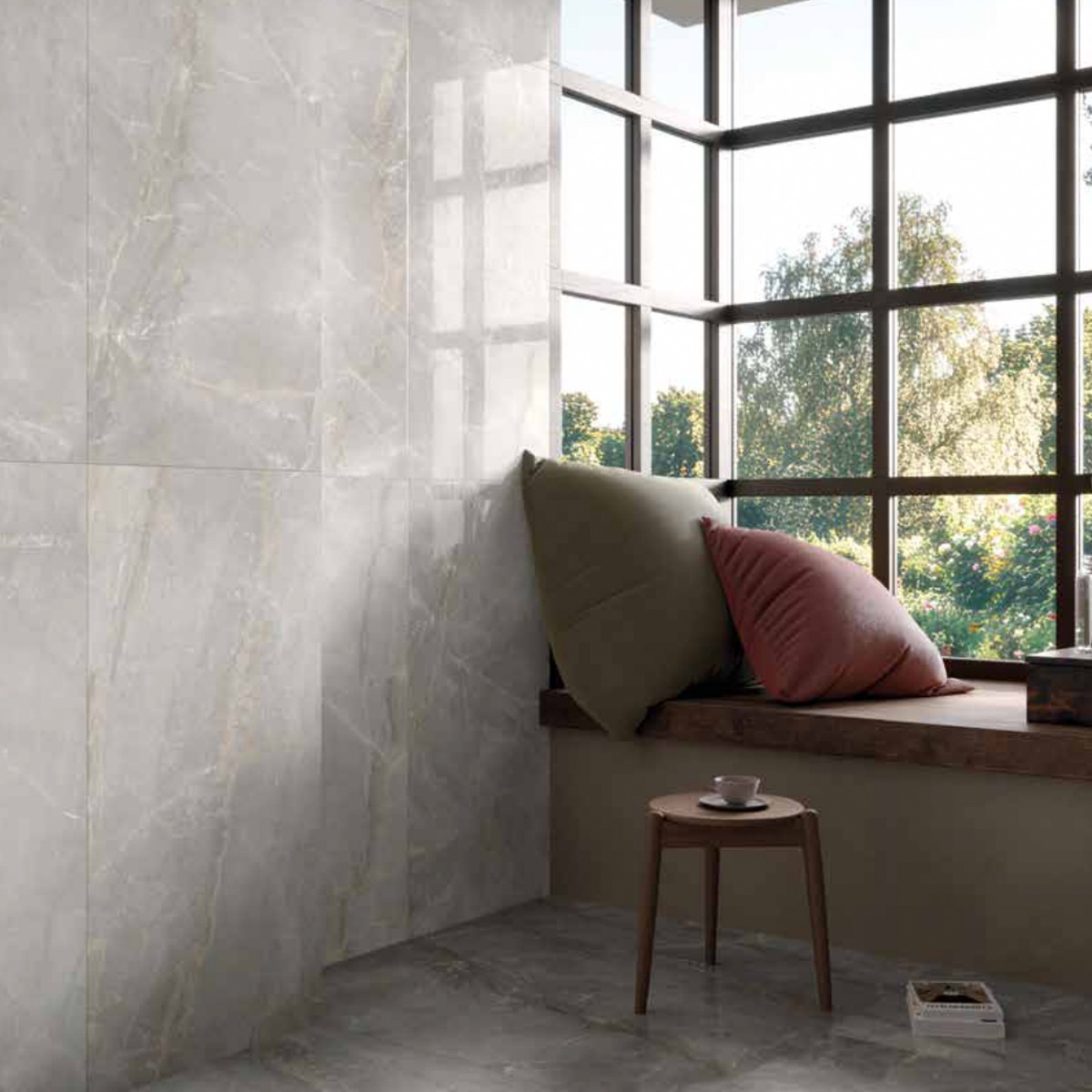 Sunny window nook with Charme - Pearl gray marble flooring and wall tiles, a wooden ledge with two large cushions, a small round wooden table, and a cup. Trees and blue sky visible outside.