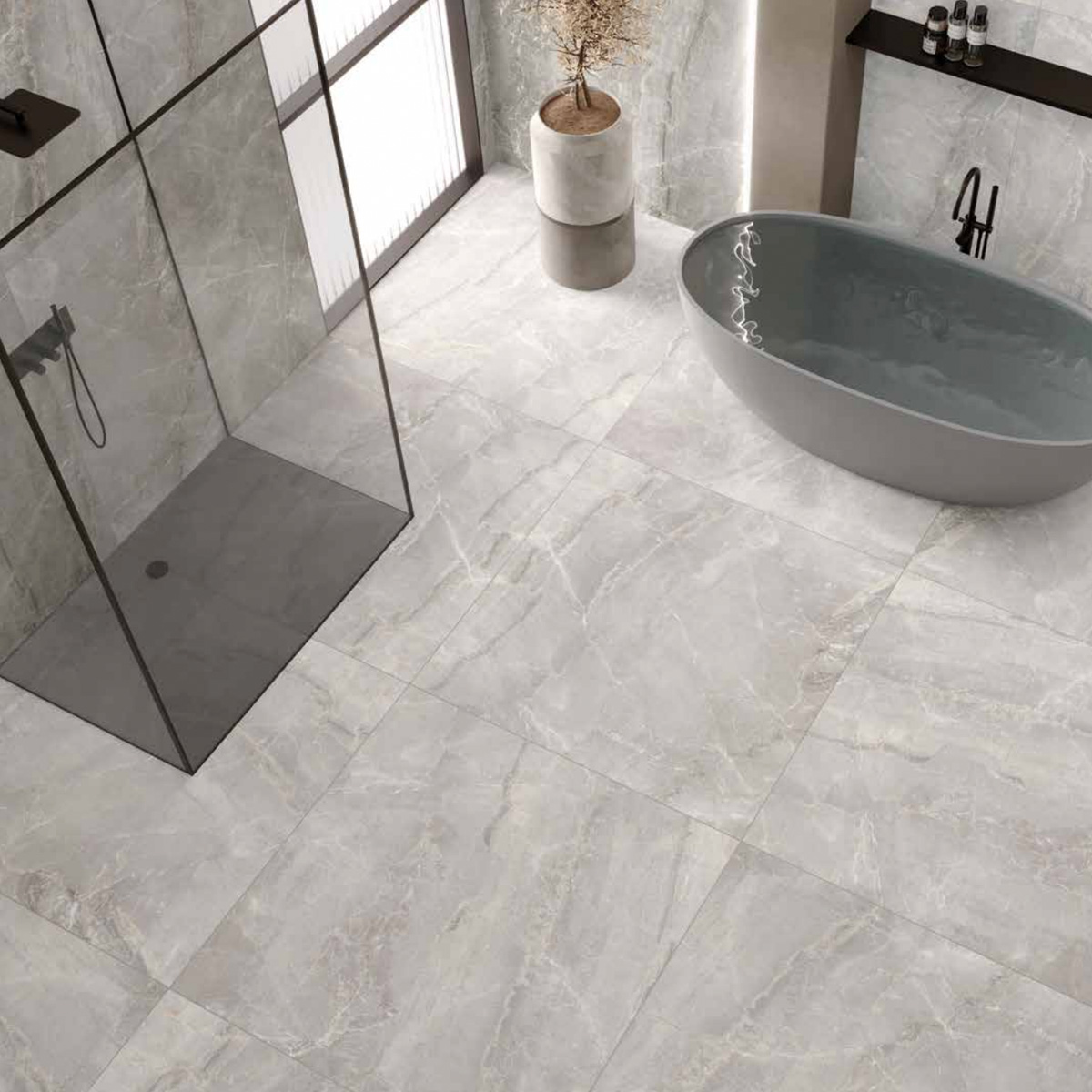 Modern bathroom featuring Charme - Pearl large gray marble tiles, a freestanding oval bathtub, a glass shower enclosure, a tall vase with branches, and a shelf adorned with decor items. Soft natural light filters in through a frosted window.