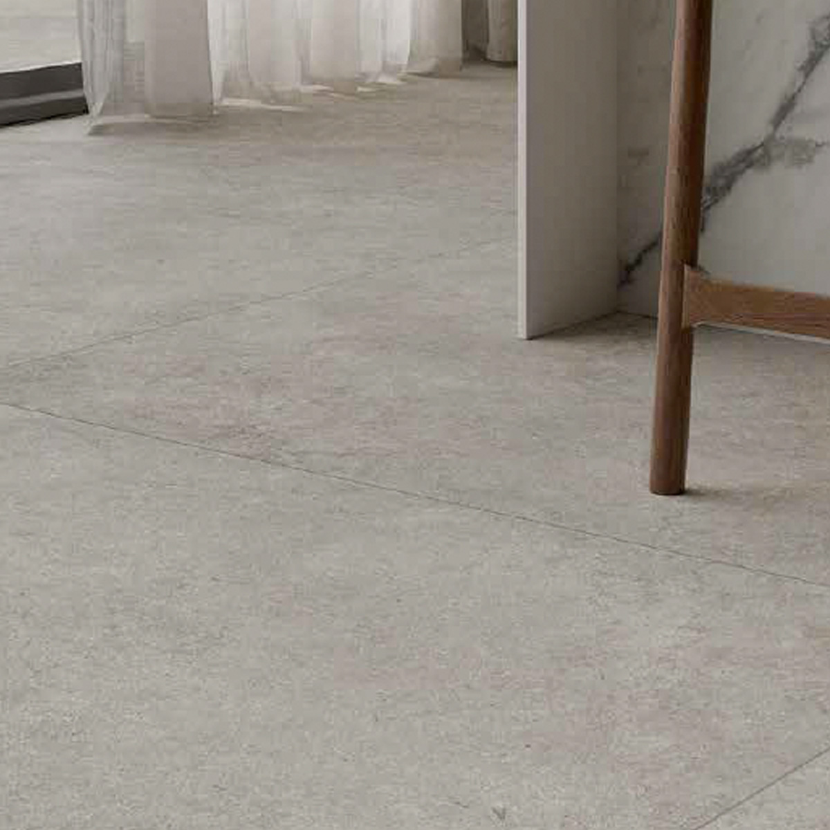 A modern, minimalist room featuring Lutezia - Grey tiled flooring with subtle lines, sheer white curtains, a glimpse of a marble-topped table, and the leg of a wooden chair.