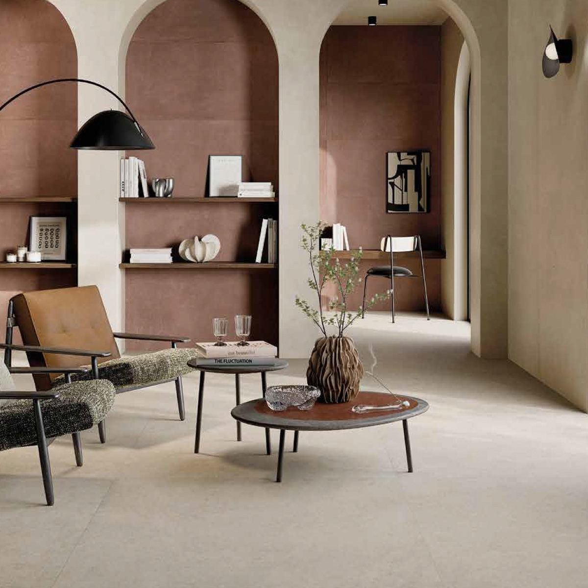 Elegant open-plan living space with large-format ivory stone-look floor tiles, arched doorways with terracotta walls, a tan leather lounge chair, two round dark coffee tables with a sculptural vase and glassware, an open shelving unit with books and decorative objects, and a large black arc floor lamp.