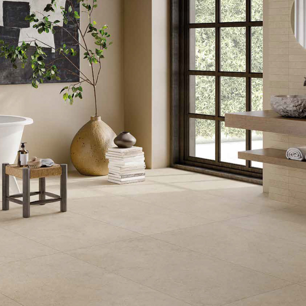 Warm contemporary bathroom with large-format taupe stone-look floor tiles and matching wall tiles, featuring a freestanding white soaking tub, a woven stool, a large textured ceramic vase with a branching plant, stacked books, a dark abstract wall painting, and a tall black-framed window overlooking greenery.