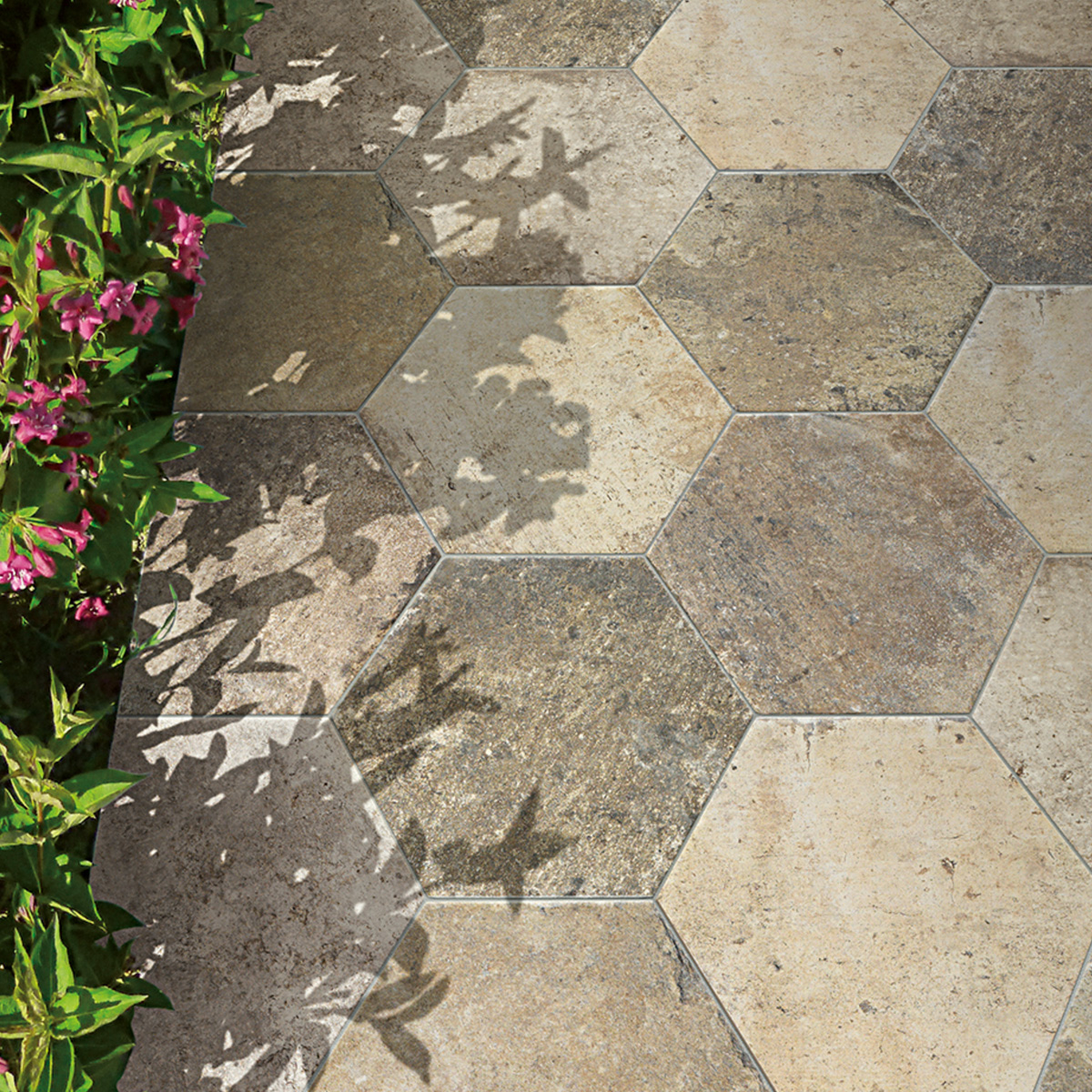 Overhead view of large hexagonal stone-look outdoor tiles in mixed sandy beige and dark gray tones, with dappled flower shadows cast across the surface and pink flowering plants visible along the left edge.