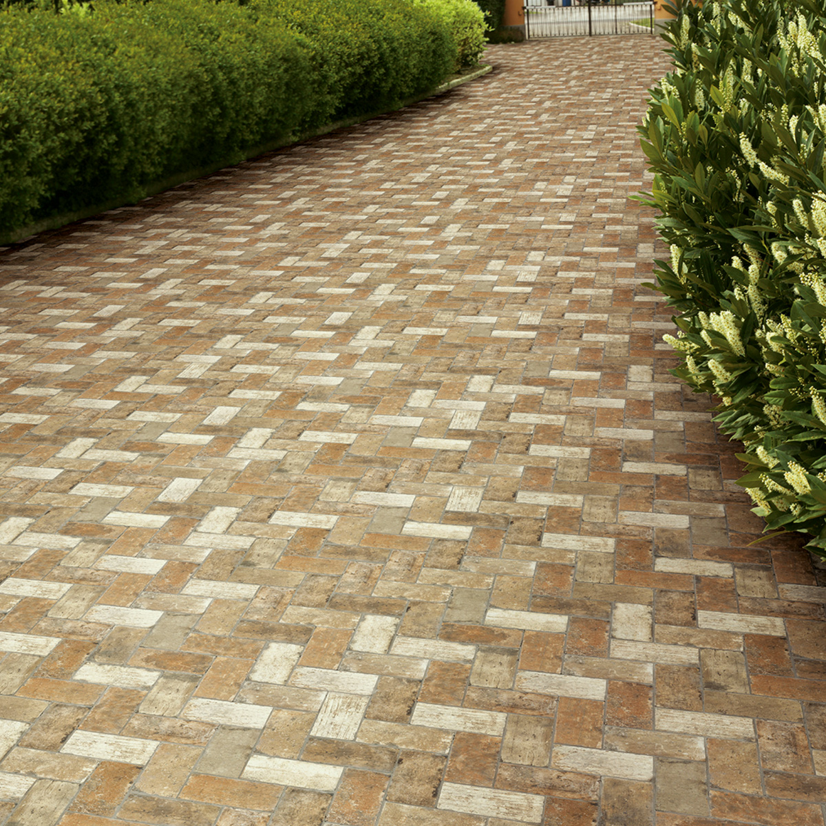 Outdoor garden pathway paved with rectangular stone-look tiles in a diagonal herringbone pattern, mixing warm rust, terracotta, and cream tones, flanked by neatly trimmed hedges and flowering shrubs with a gated entry visible in the distance.