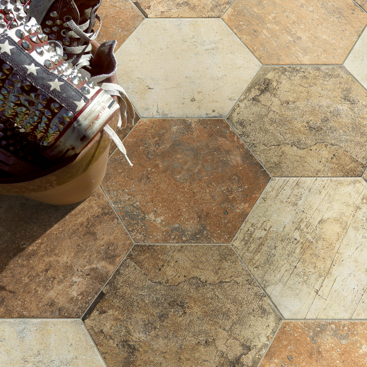 Overhead view of large hexagonal stone-look floor tiles in warm rust, terracotta, and sandy beige tones with natural variation and weathered texture, with a decorative ceramic pot and studded sneaker visible in the upper left corner.