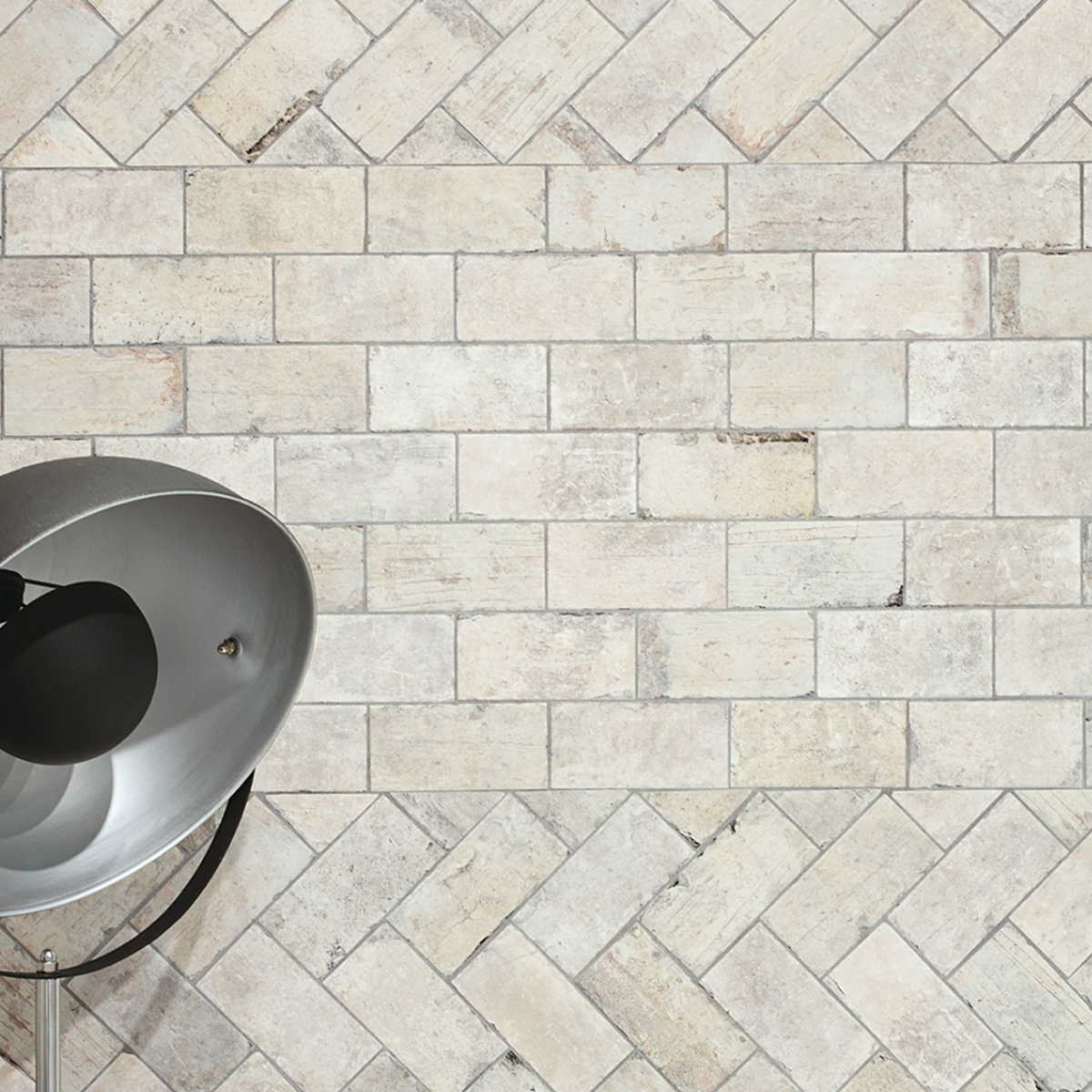 Overhead view of cream and off-white stone-look rectangular tiles shown in two laying patterns: a straight horizontal brick layout in the center and diagonal herringbone at the top and bottom, with a silver industrial floor lamp visible in the lower left.
