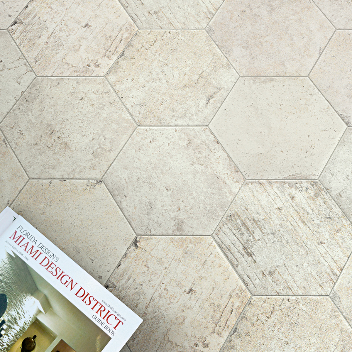 Overhead view of large hexagonal stone-look floor tiles in cream and warm beige tones with subtle veining, with a design magazine partially visible in the lower left corner.