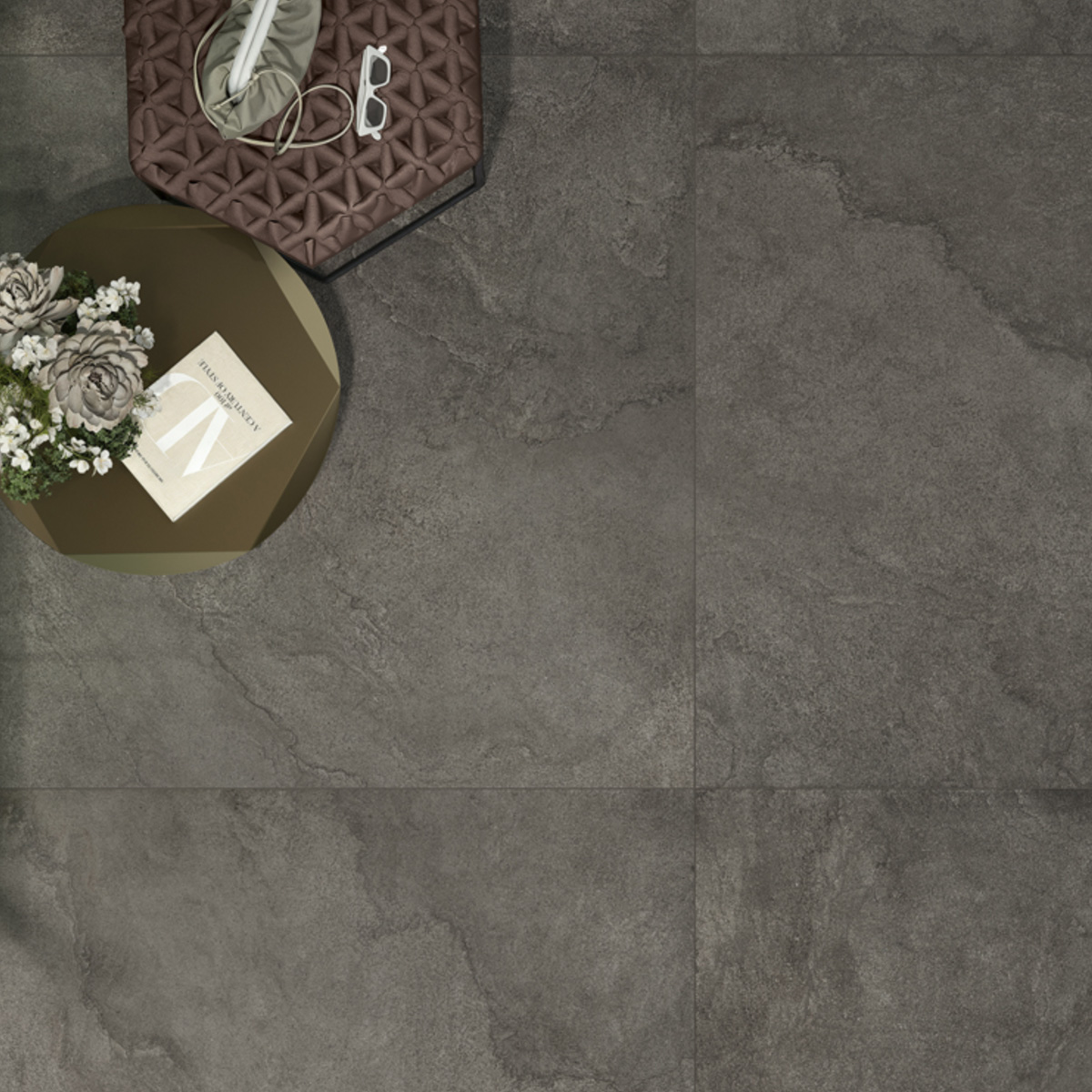 A round table adorned with flowers and a book is placed beside a quilted brown cushion and a bag on the Pulse - Shadow dark grey tiled floor.