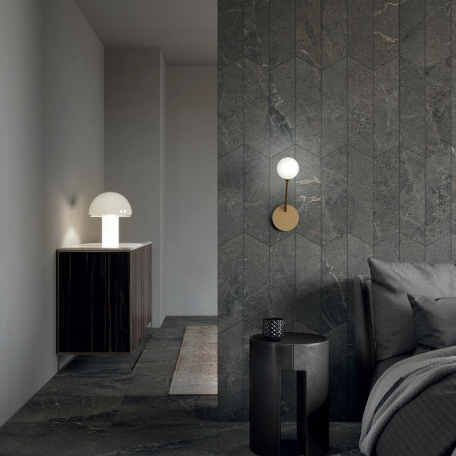 A modern bedroom featuring Anthology/Anthea - Dark stone walls and floors, a gray bed, a small round side table, a wall-mounted lamp, and a white table lamp on a cabinet in the hallway.
