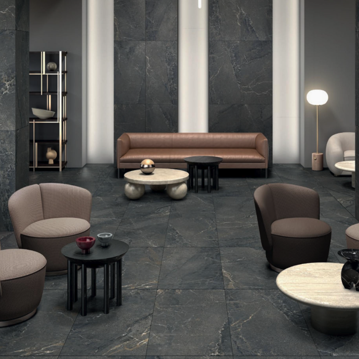 A modern lounge featuring Anthology/Anthea - Dark marble floors and walls, complemented by neutral armchairs, a brown leather sofa, round coffee tables, a tall shelf with décor, and a standing lamp, all under soft, minimalist lighting.