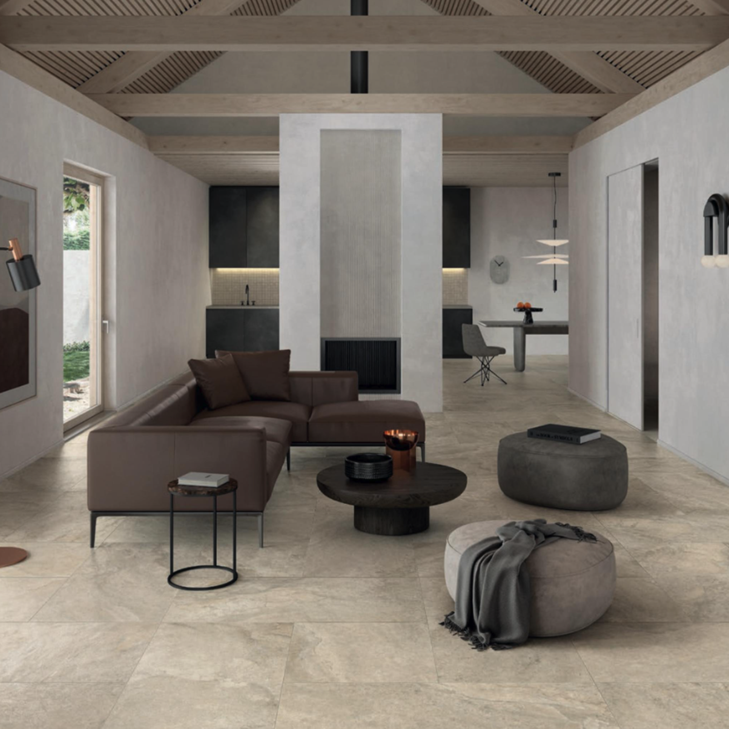 Modern living room with neutral tones, featuring the Anthology/Anthea - Desert sectional sofa, round coffee tables, poufs, and an open kitchen in the background. Large windows let in natural light, while wooden ceiling beams add warmth to the minimalist decor.