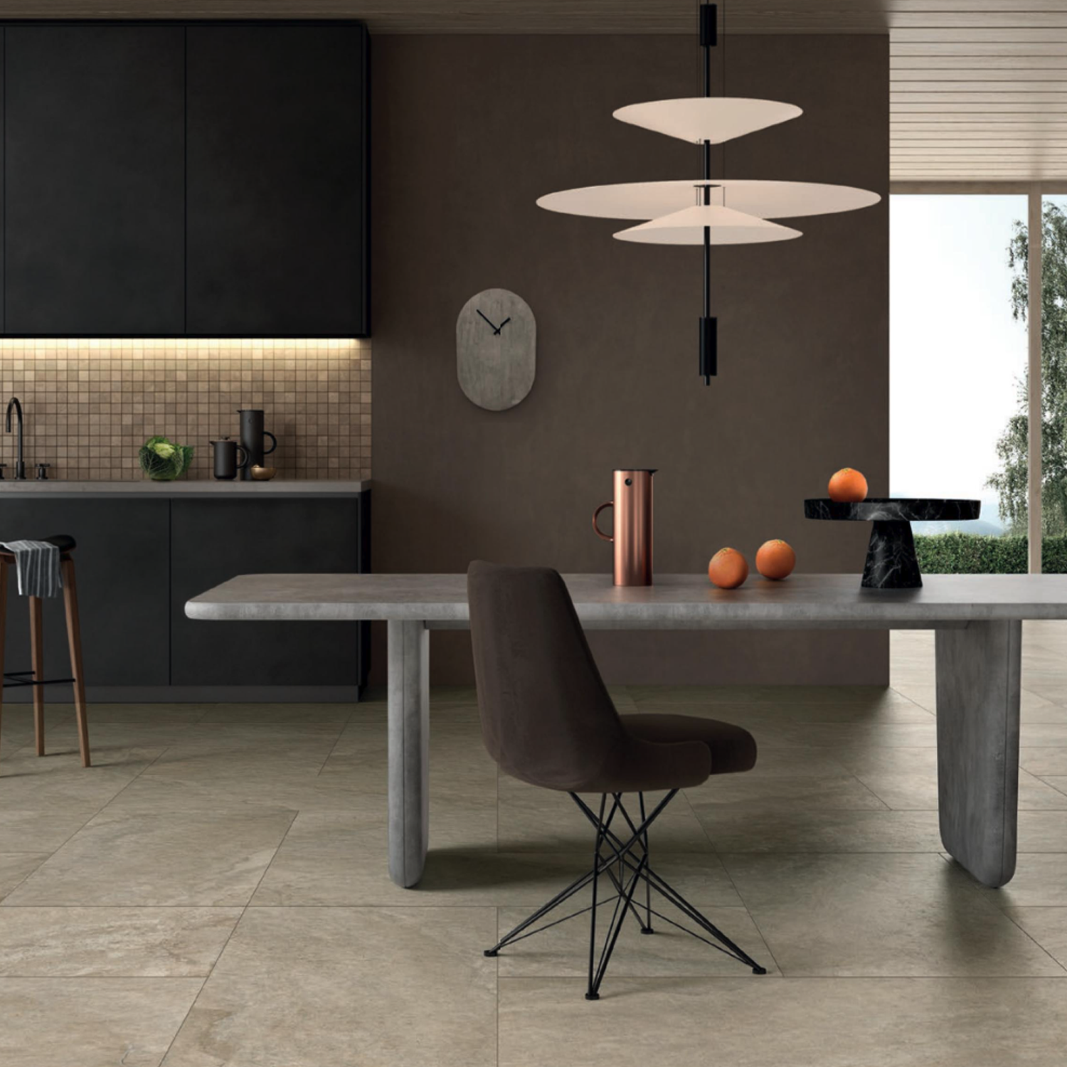 Modern kitchen featuring large Anthology/Anthea - Desert tiles, a sleek dining table, brown chair, pendant lights, fruit on the table, dark cabinets, and a wall clock. Neutral tones and the distinctive tile design contribute to a minimalist, contemporary look.