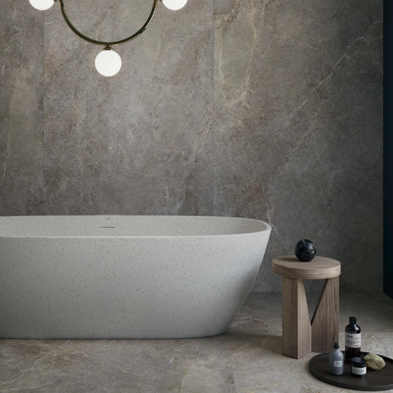 Modern bathroom featuring Anthology/Anthea - Earth gray marble tiles on the walls and floor, with a freestanding white tub, round wooden stool displaying bath products, and a circular hanging light fixture.