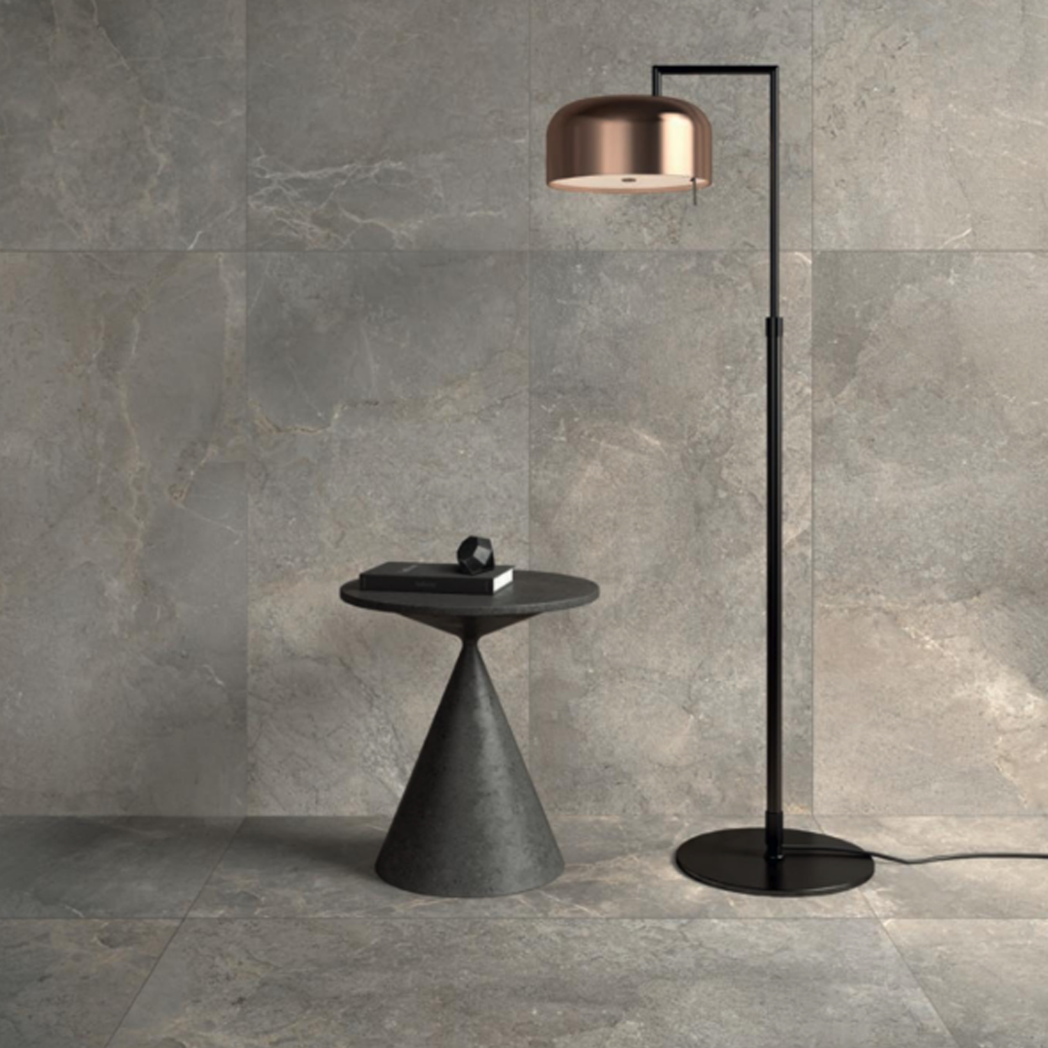 An Anthology/Anthea - Earth side table styled with a book and decor is placed next to a sleek black floor lamp with a copper shade, all set against an Anthology/Anthea - Earth gray stone-tiled wall and floor.