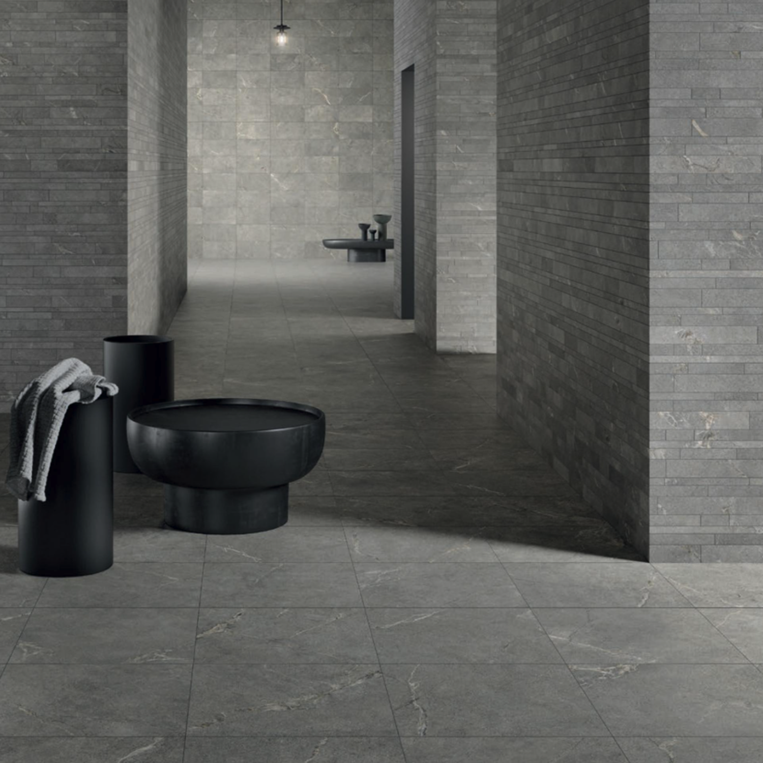 A modern, minimalist room features Anthology/Anthea - Gray textured walls and floor, a black basin, a cylindrical stand with a towel, and dim ceiling lighting, creating a sleek industrial aesthetic.