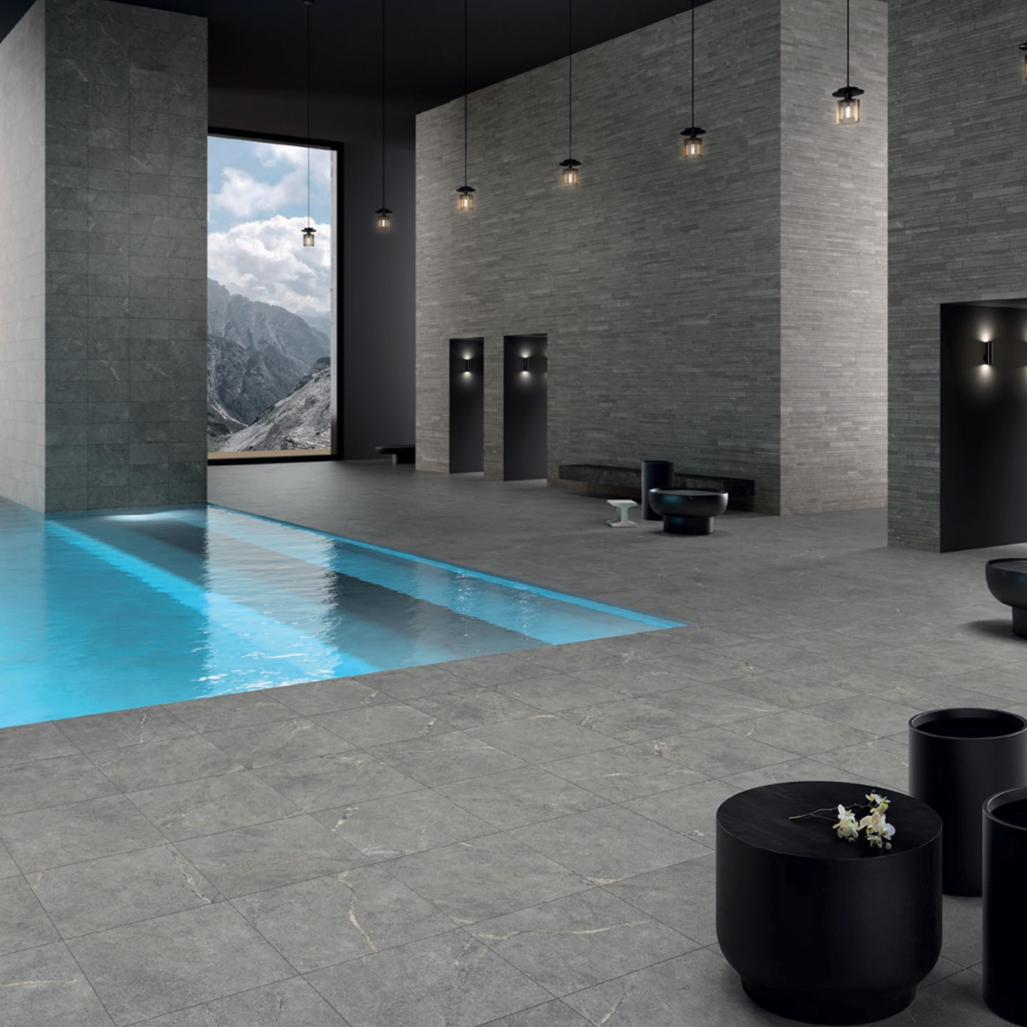 Indoor pool featuring Anthology/Anthea - Gray stone tiles, complemented by modern black furniture, wall lights, and large windows revealing a mountain view under a cloudy sky.