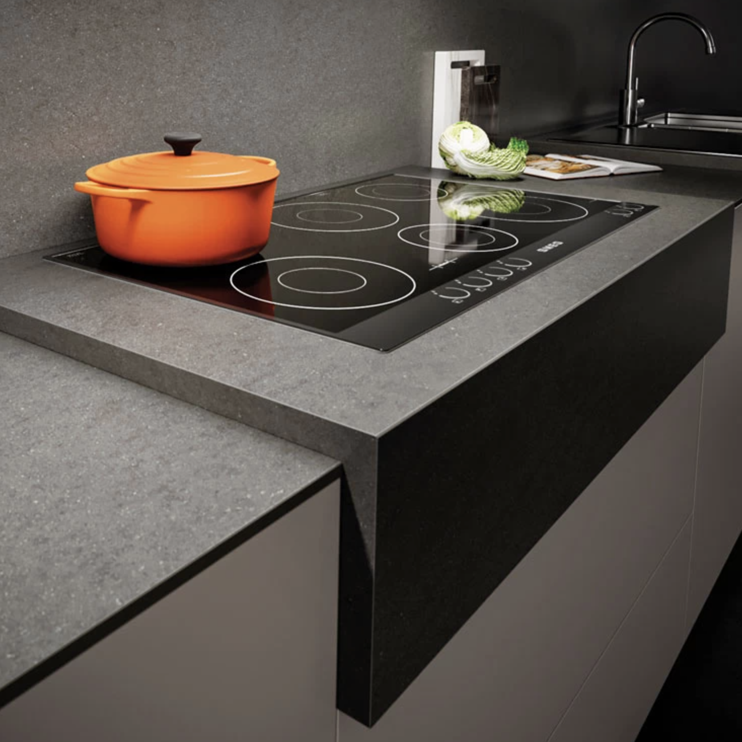 A modern kitchen showcases a Basaltina Volcano gray countertop with a black electric cooktop. An orange pot sits on the burner, while a sink with a faucet is in the background. Utensils and a decorative item are arranged on the counter.