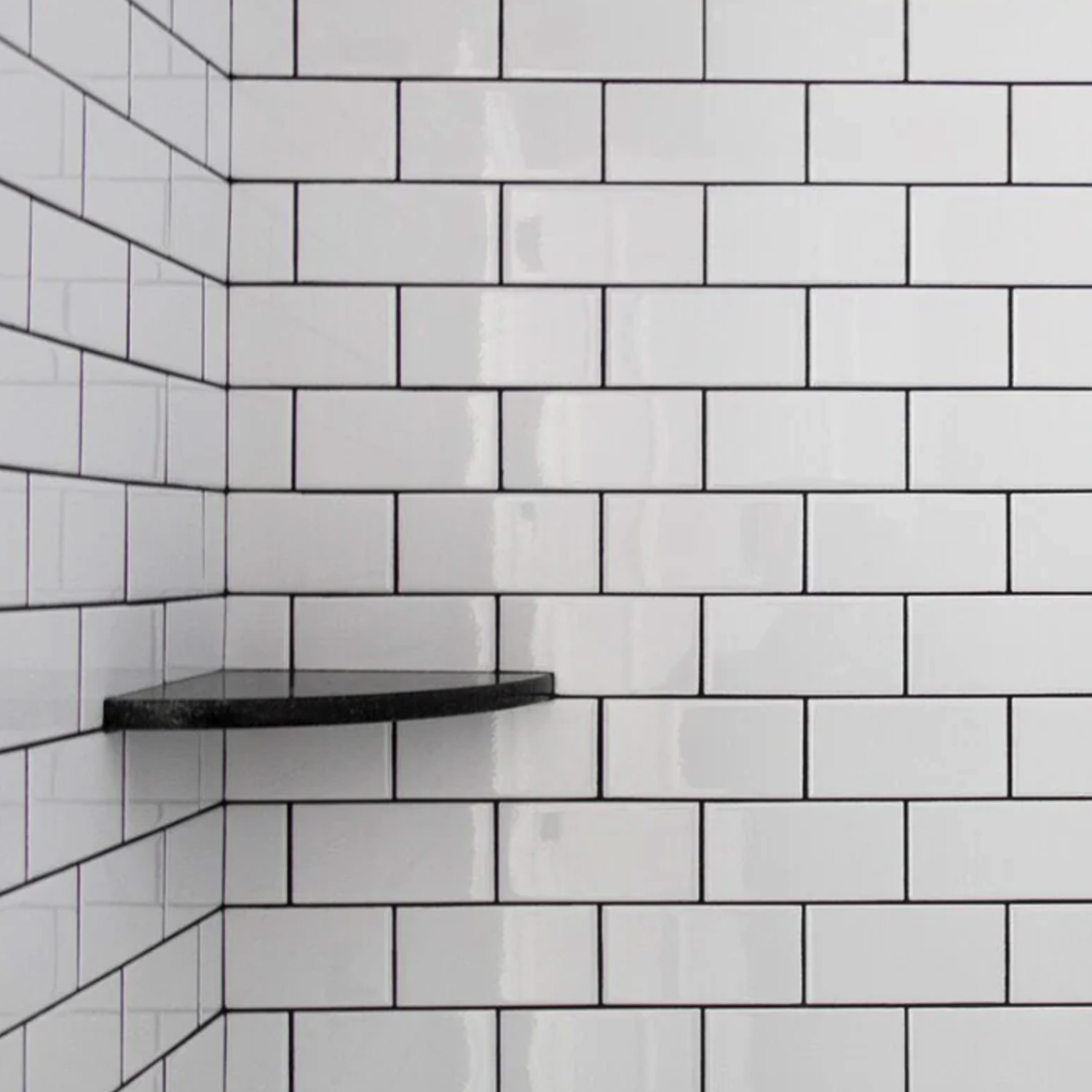 Glossy white subway tiles with black grout are arranged in a staggered brick pattern on the shower wall. The left side features a single Bath Accessories (Corner Shelf) - Black mounted in the corner.