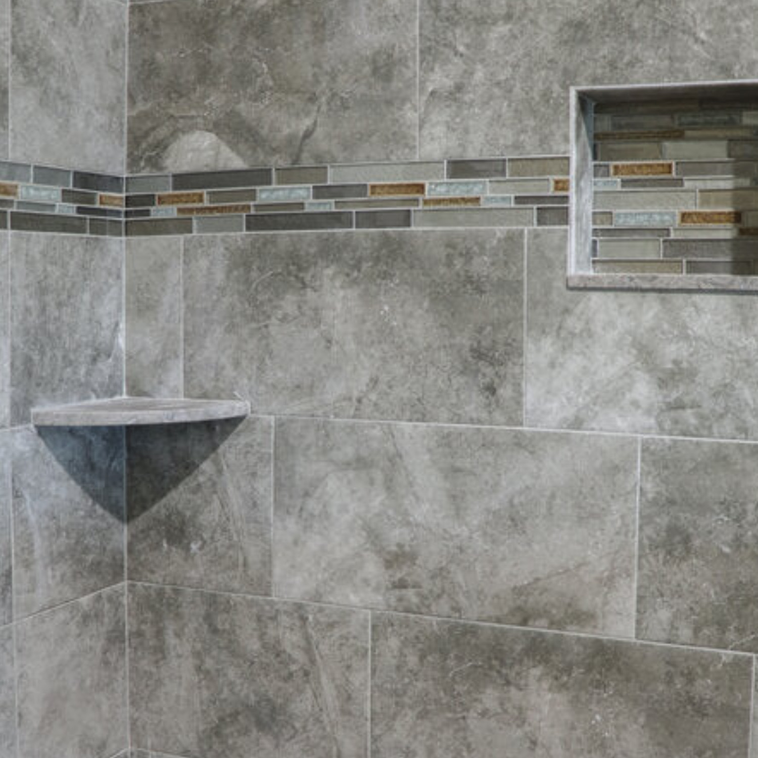Shower wall featuring large gray tiles, a horizontal accent strip of small multicolored tiles, a built-in Sage Gray corner shelf from the Bath Accessories collection, and a recessed niche lined with matching accent tiles.