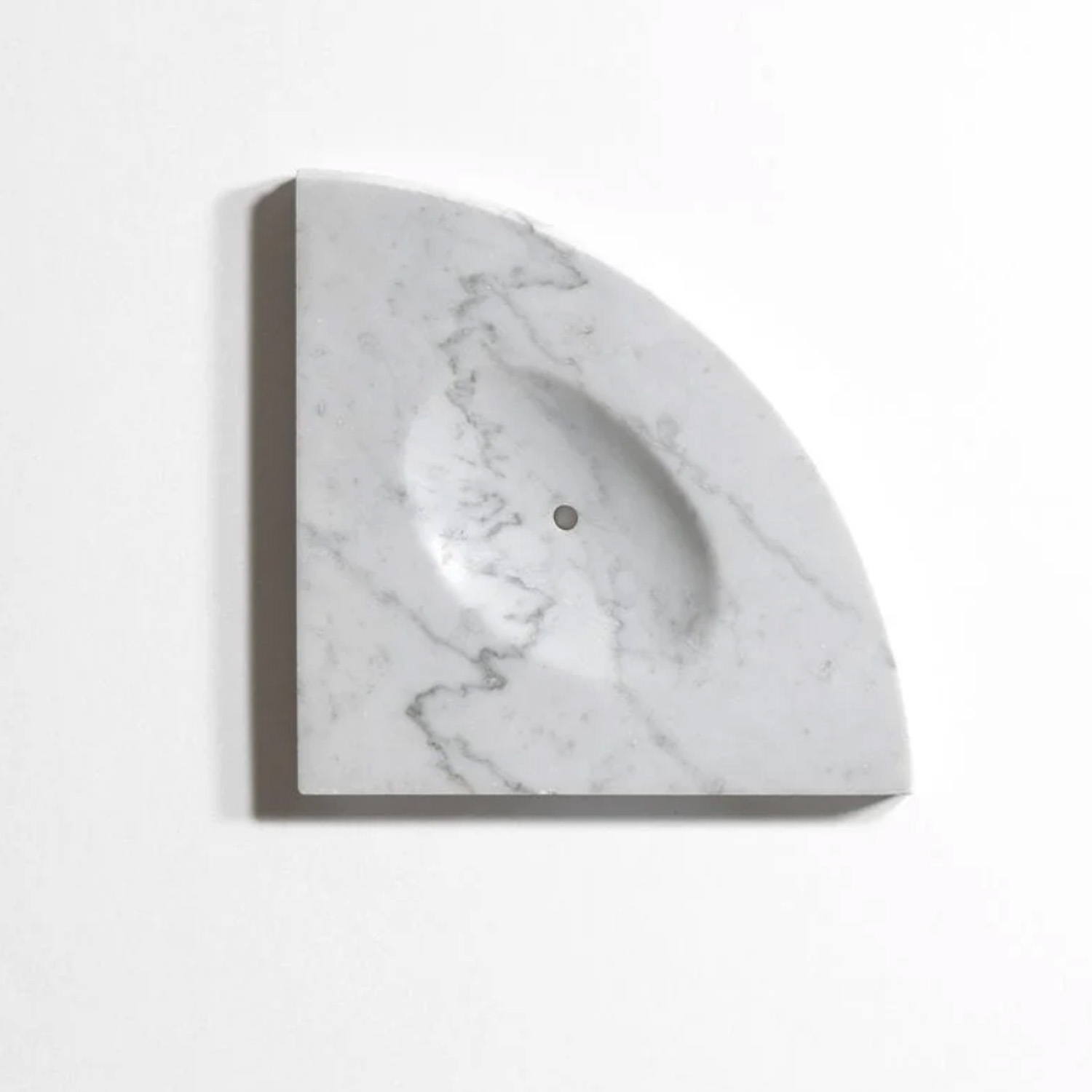 A Bath Accessories (Corner Shelf) - Venatino Carrara, crafted from marble with subtle gray veining, features a quarter-circle shape with a smooth, shallow indentation and a small hole at the center. It is mounted on a white wall.