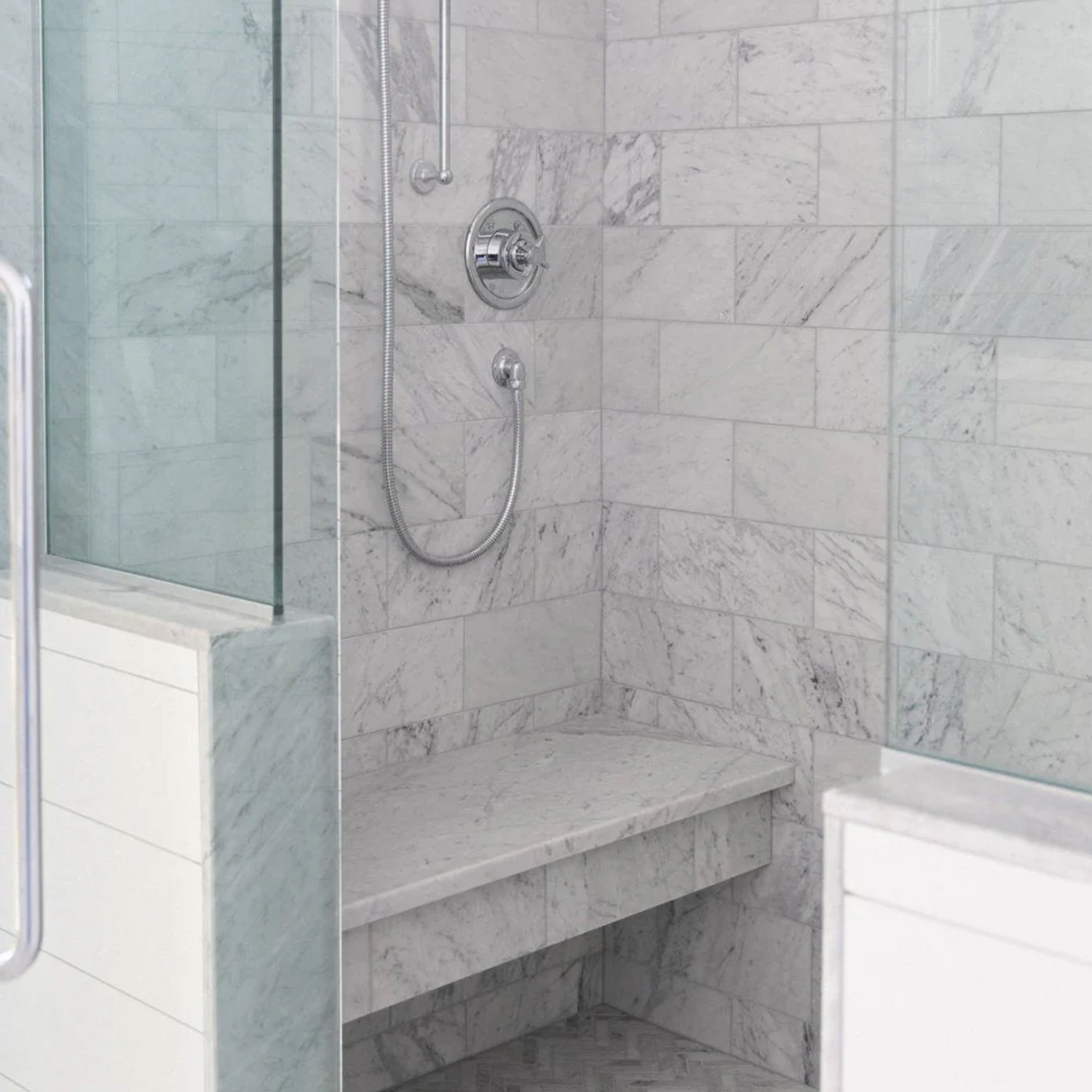 A modern shower featuring a built-in Bench - Colorado Calacatta, grayish-white marble tiles, a glass door, and a silver showerhead with a handheld attachment.