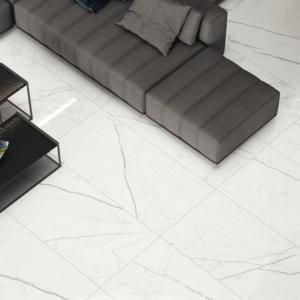 A sectional gray sofa with cushions sits beside two black metal coffee tables on a Beyond Calacatta – Classic White floor with gray marble veining.