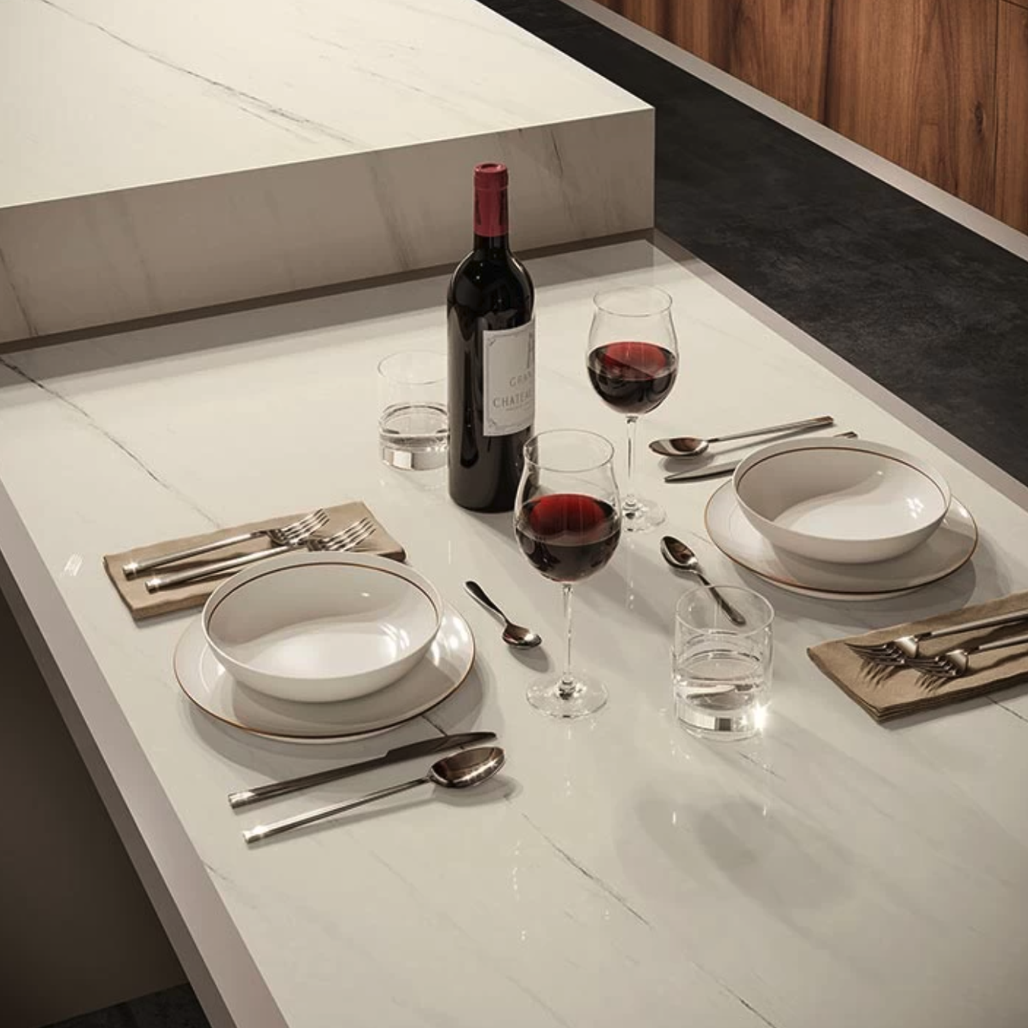 A modern table set for two with white dishes, cutlery, glasses of water, red wine, and a bottle of wine rests on a Bianco Dolomite A countertop. Warm wooden paneling can be seen in the background.