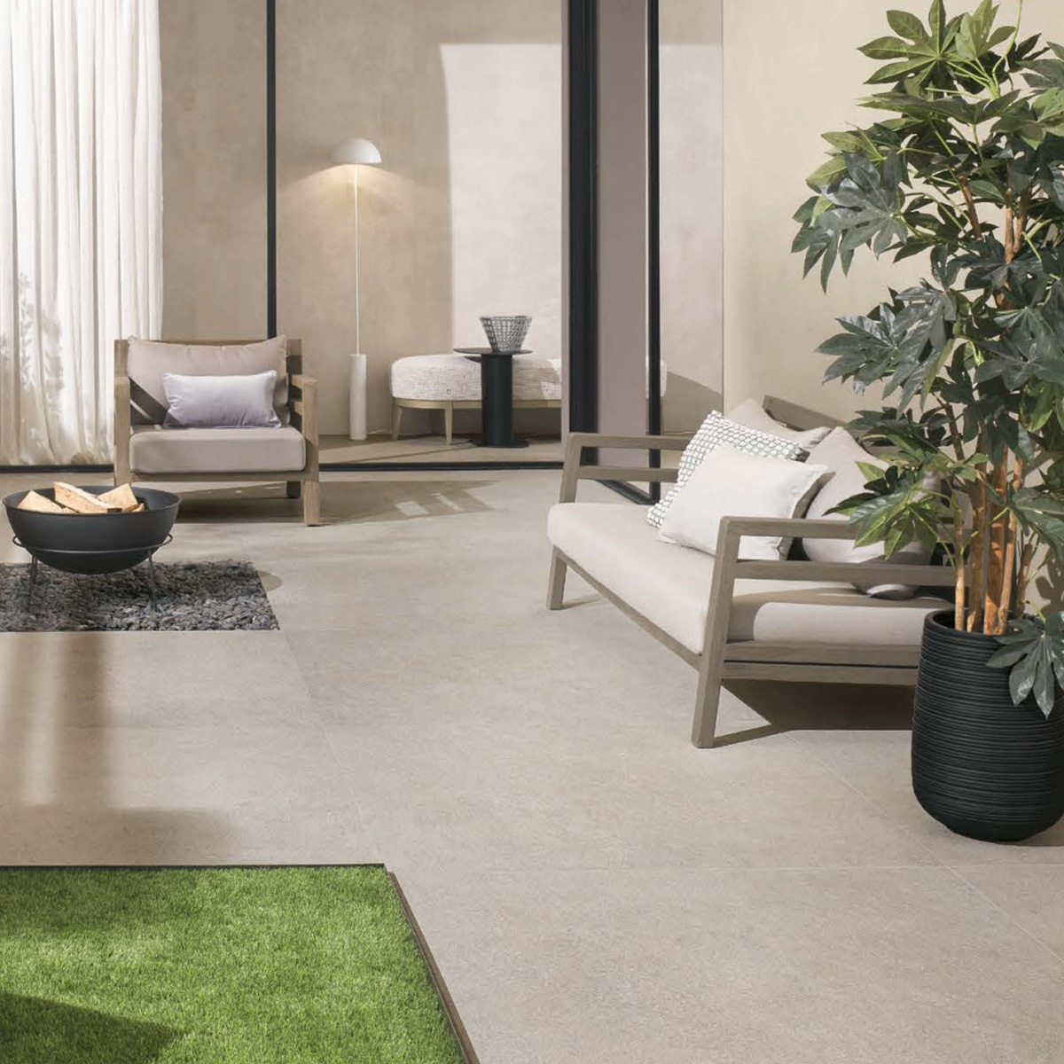 Modern living room with beige walls, light wood furniture, white and gray pillows, a potted plant, floor lamp, curtains, and a bowl-shaped centerpiece on a small dark rug featuring Boost Mineral - Grey. A green rug is partially visible in the foreground.