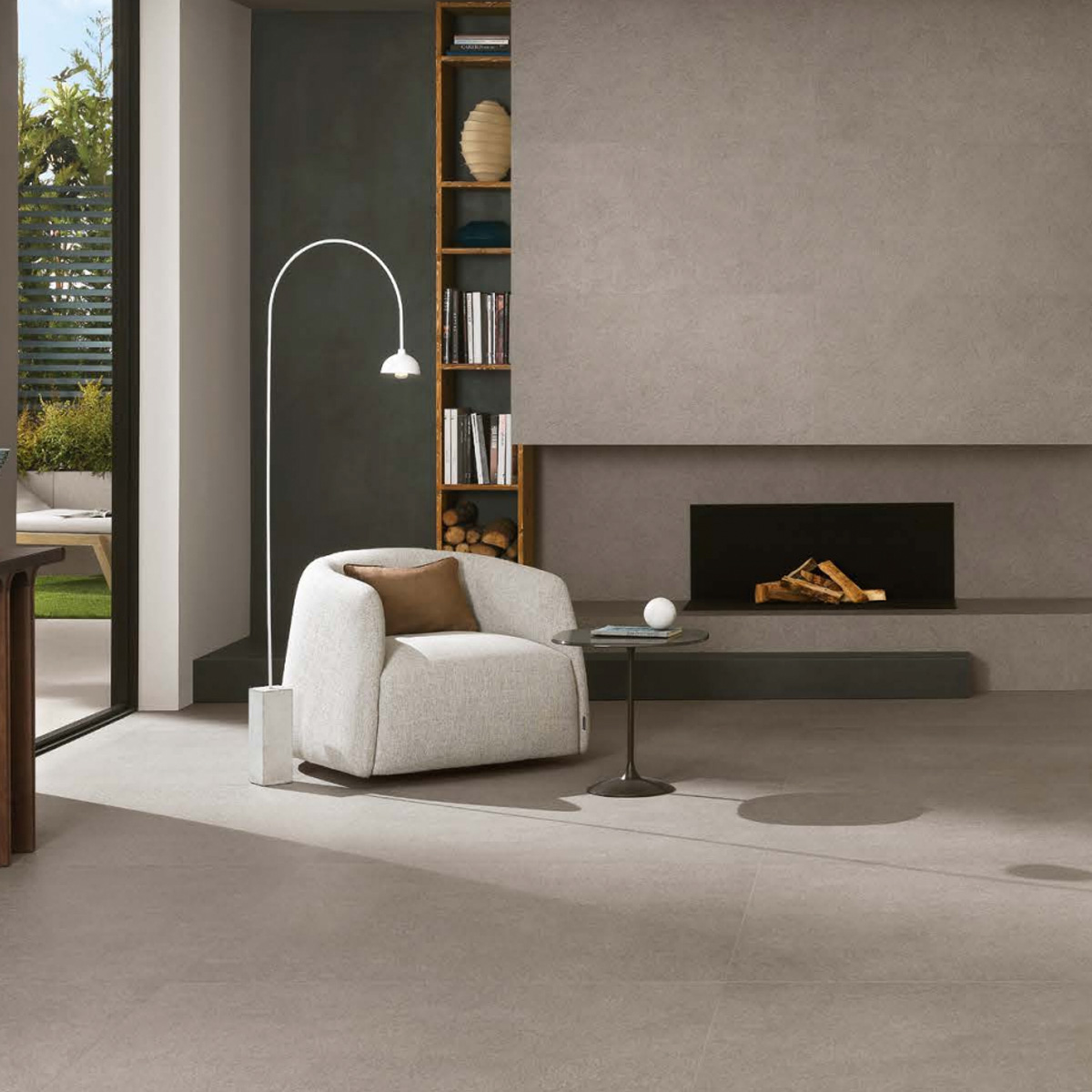 A modern living room featuring the Boost Mineral - Grey armchair, paired with a small round table, a floor lamp, built-in shelves filled with books, a cozy fireplace, and large windows that flood the space with natural light.