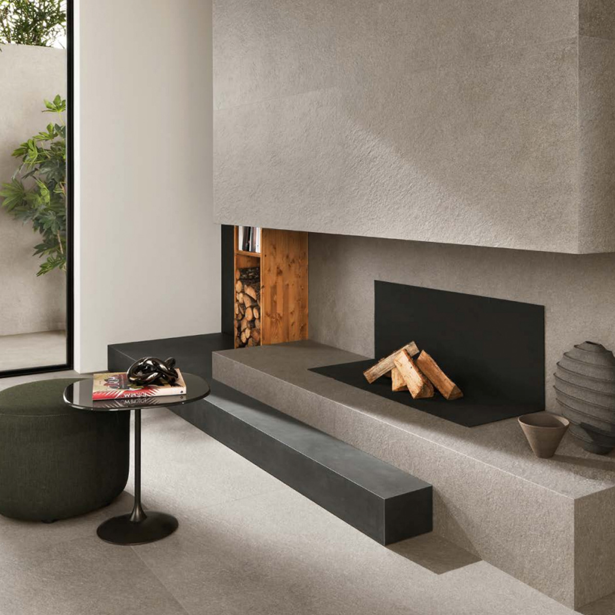 Modern living room featuring a minimalist Boost Mineral - Grey stone fireplace, stacked firewood, a round black table with books and decor, green ottoman, and a plant visible through a glass door.