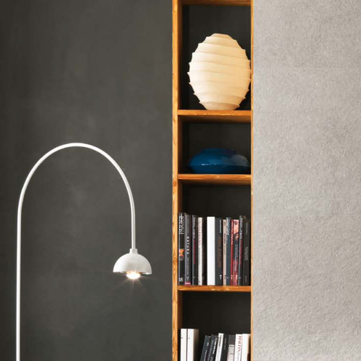 A white floor lamp stands beside a wooden bookshelf built into a wall, holding books and decorative items including a white sculptural vase and a blue bowl. The background features wall sections in Boost Mineral - Tarmac.