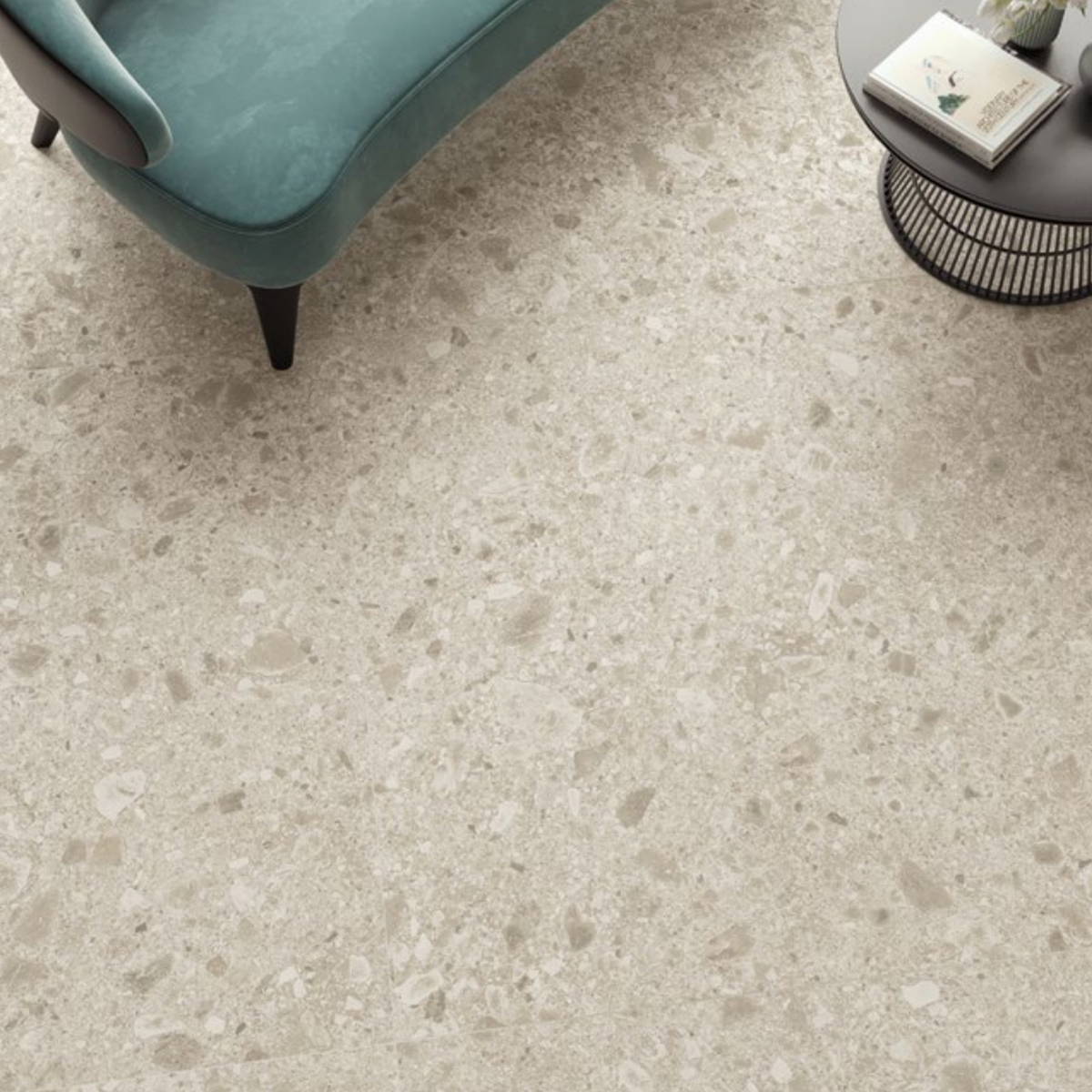 A Boost Mix - Ivory terrazzo floor with scattered stone pieces is visible, along with part of a teal upholstered sofa and a round black metal side table holding a book and a small plant.
