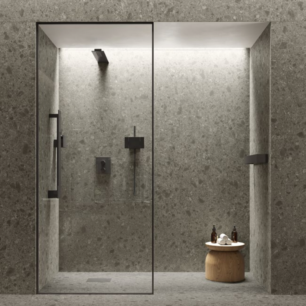 Minimalist shower featuring Boost Mix - Smoke gray stone tiles on the walls and floor, glass door, black fixtures, and a small round wooden stool just outside holding bottles and a towel. The space is softly illuminated from above.