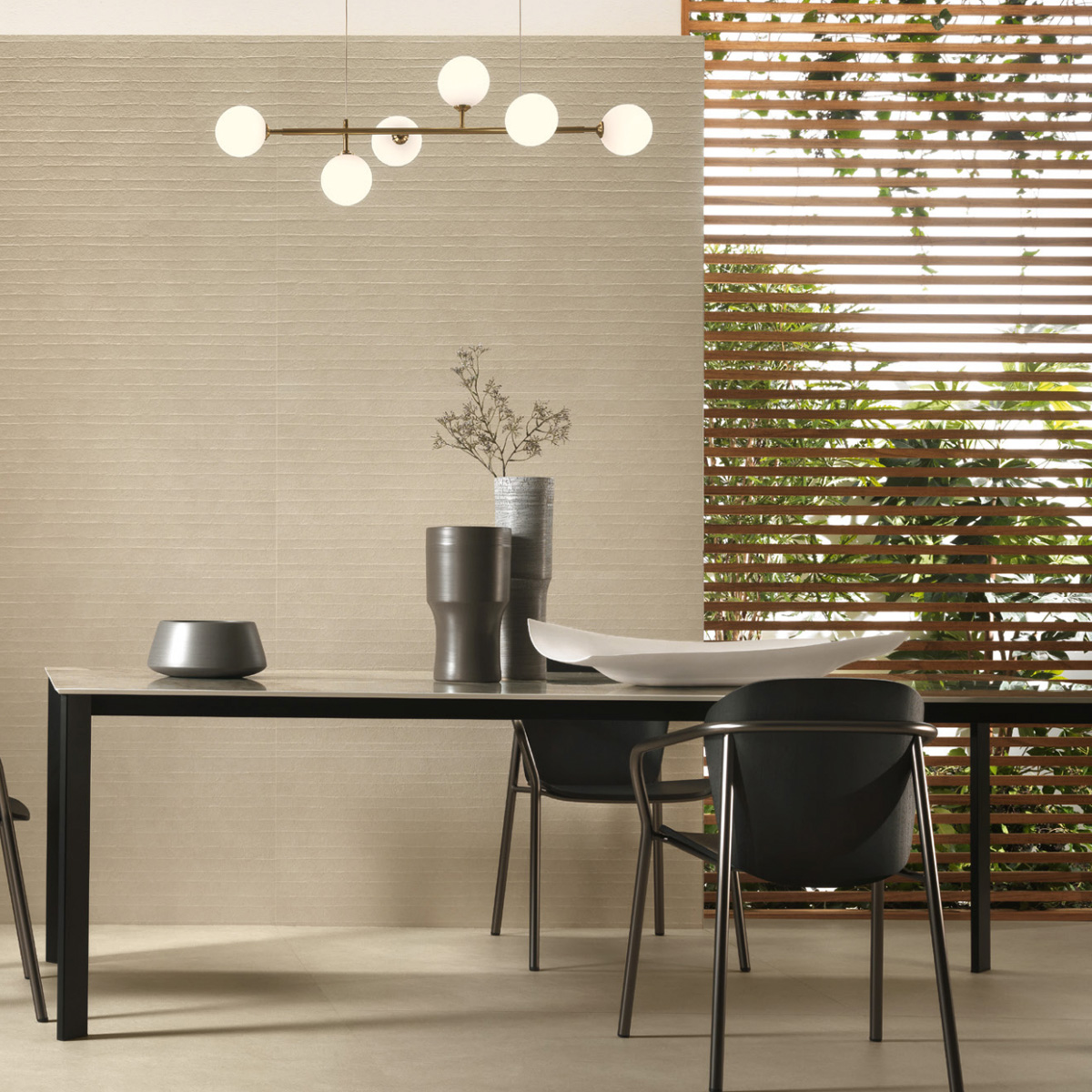 Modern dining room featuring the Boost Natural Pro - Oat table, paired with dark chairs, minimalist vases, and a white bowl. Wooden slat wall opens to outdoor greenery, while a contemporary light fixture with round bulbs illuminates the space. The design showcases a neutral color palette and clean lines.