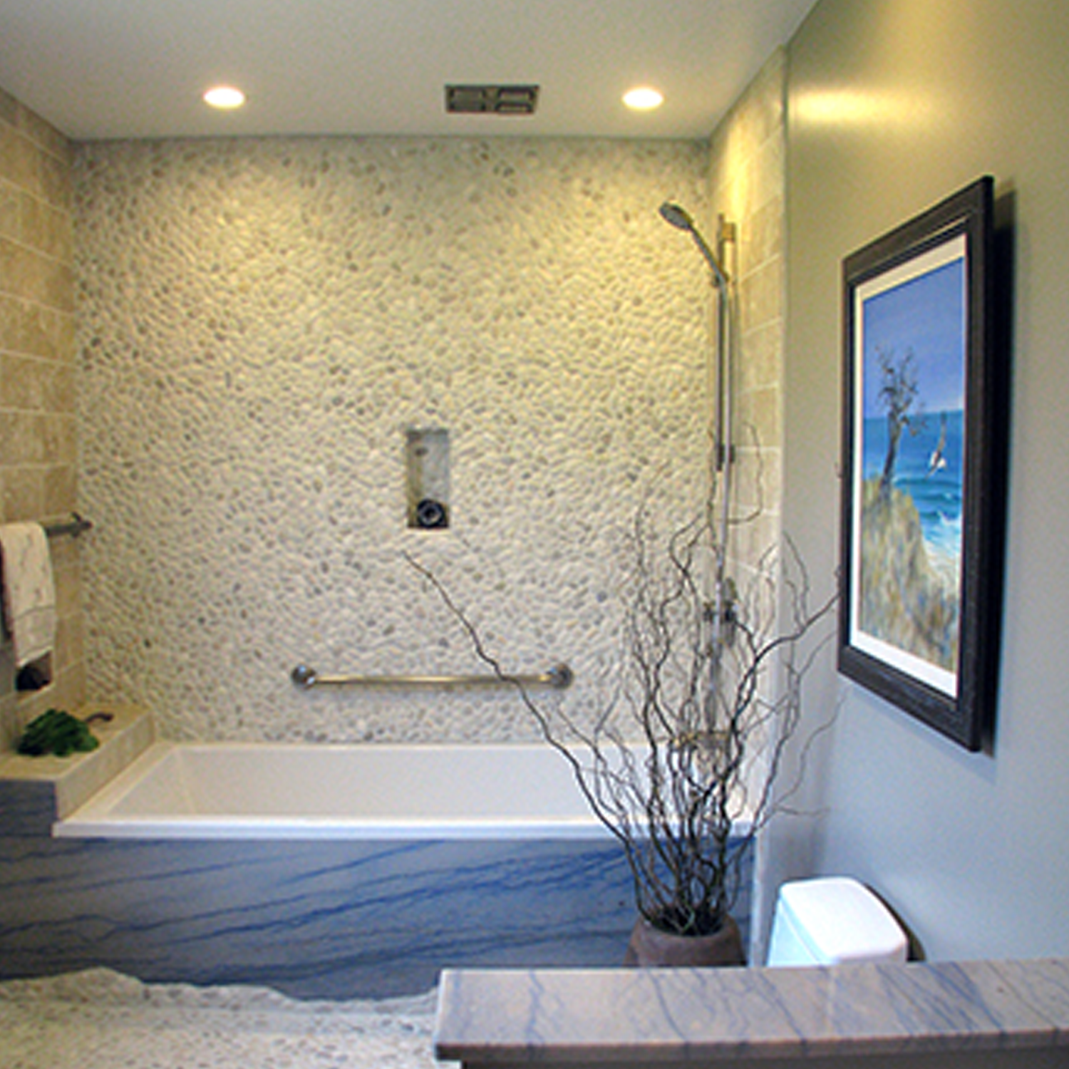 Modern bathroom featuring a Botany Bay - Cream Sliced textured stone wall, bathtub with metal grab bar, showerhead, wall art, tall vase with branches, white toilet, and soft lighting from ceiling spotlights.