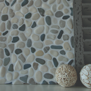 Botany Bay - Shadow Blend, featuring round white and blue pebbles embedded in the surface, is displayed next to a dark shutter and two decorative, textured cream balls on a gray tabletop.