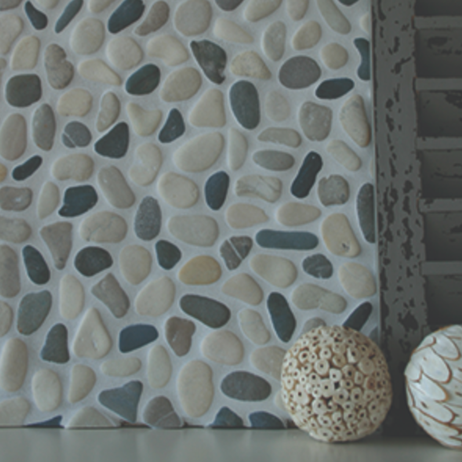 Botany Bay - Shadow Blend, featuring round white and blue pebbles embedded in the surface, is displayed next to a dark shutter and two decorative, textured cream balls on a gray tabletop.