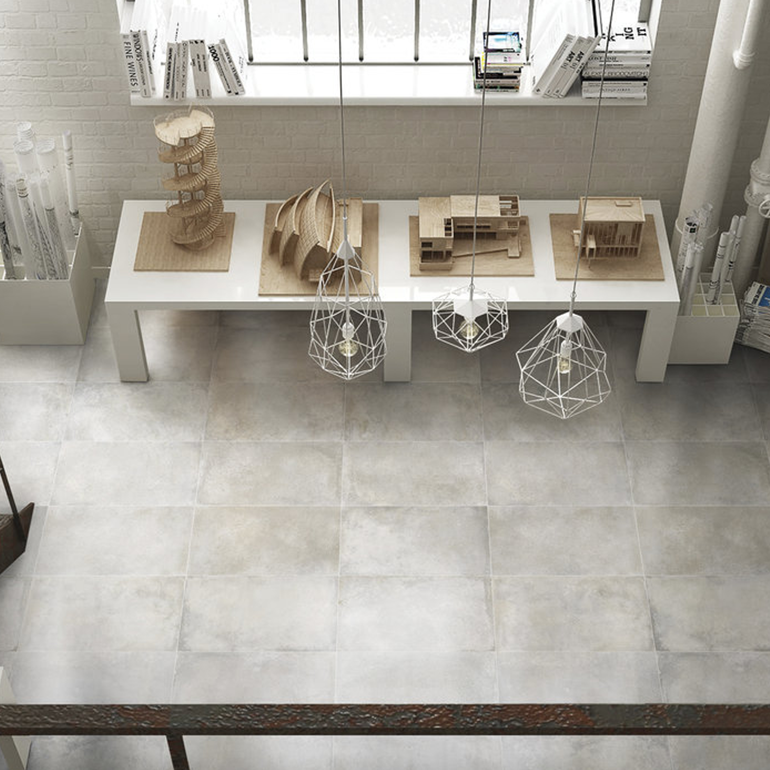 A bright room with large Brooklyn - Cemento Argent floor tiles, a white table holding three architectural models, hanging geometric pendant lights, books on the window sill, and rolls of paper in the corners.
