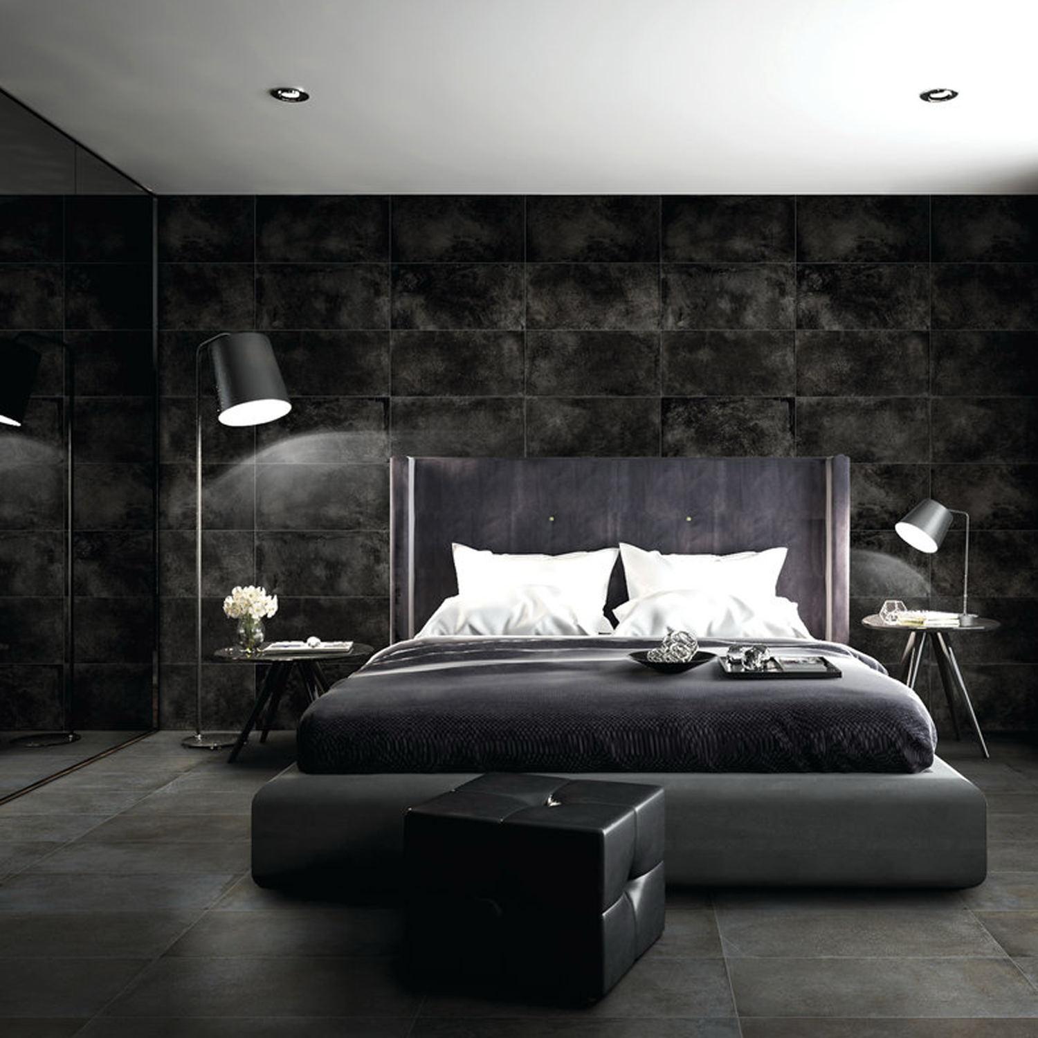Minimalistic and sophisticated modern bedroom featuring a striking accent wall with Brooklyn - Cemento Black tiles, complemented by a gray bed with white pillows, two floor lamps, decorated side tables, and a black ottoman at the foot of the bed.