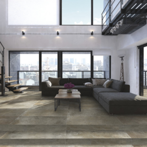 A modern loft living room featuring large windows, a dark sectional sofa, and a coffee table with flowers is anchored by the Brooklyn - Cemento Toupe flooring. An open staircase leads upward, and city buildings are visible outside.
