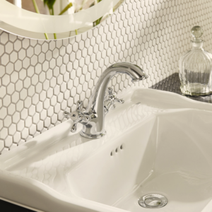 Close-up of a white sink with a chrome faucet and CC Mosaics - White hexagonal tile backsplash. A clear glass bottle sits on the counter beside the sink, and a round mirror is partially visible above.