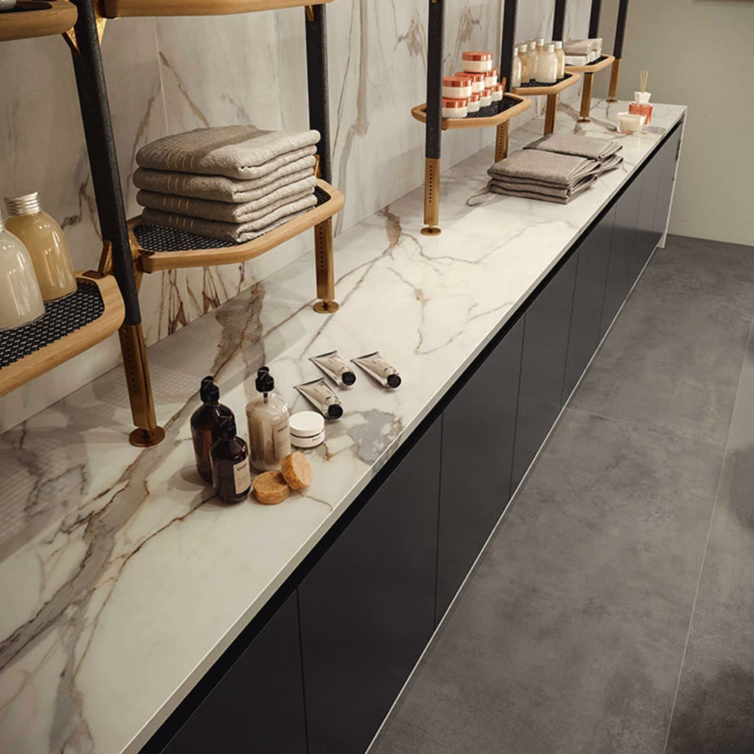 A modern bathroom features a Calacatta Imperiale marble counter, neatly folded towels, toiletries arranged in bottles and jars, and shelves above stocked with additional towels and bathroom essentials. The space is completed with a gray floor and black cabinets.