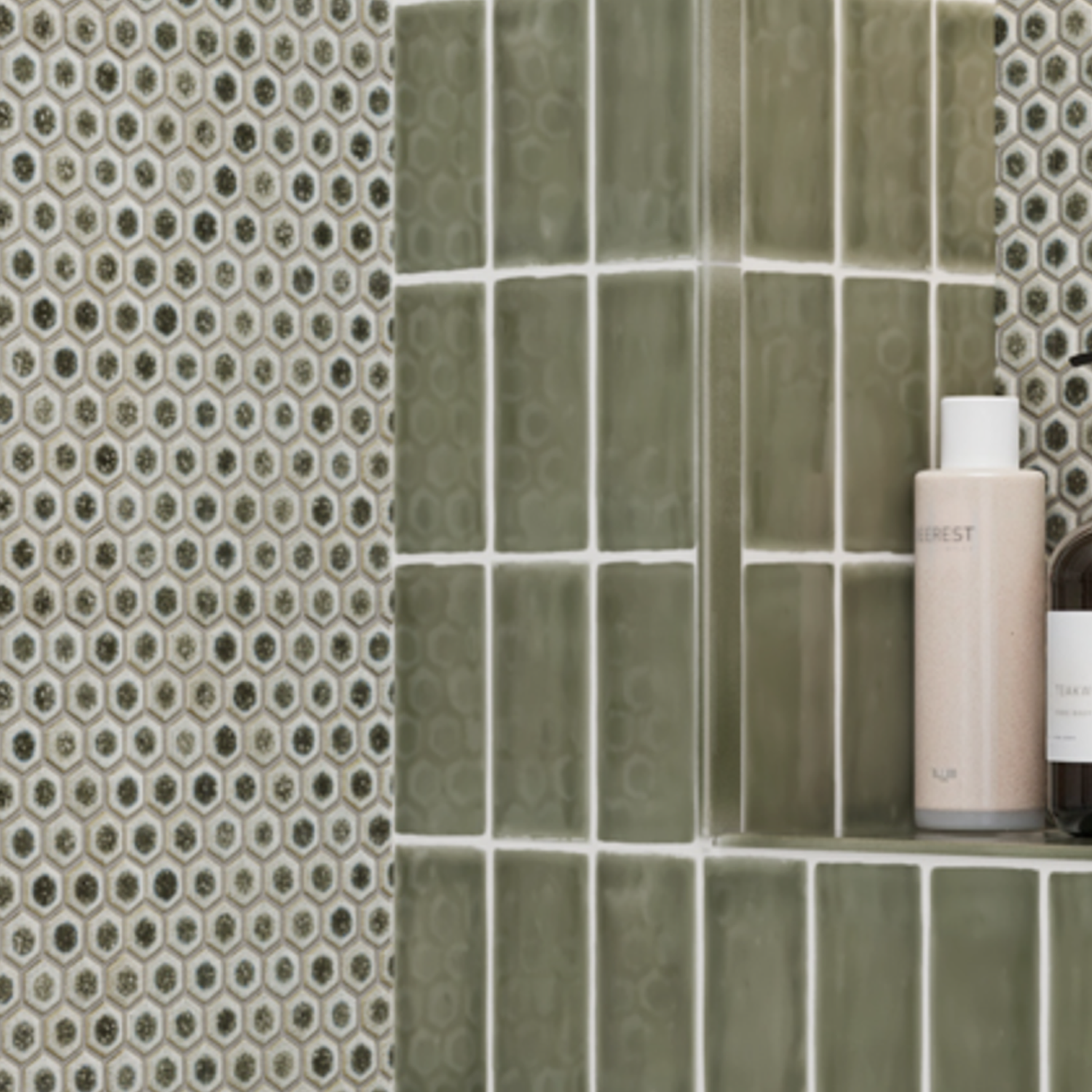 Close-up of a shower shelf featuring Chameleon - Sage Green square tiles, hexagonal mosaic wall tiles, and two bottles—one cream-colored and one dark—arranged on the shelf.